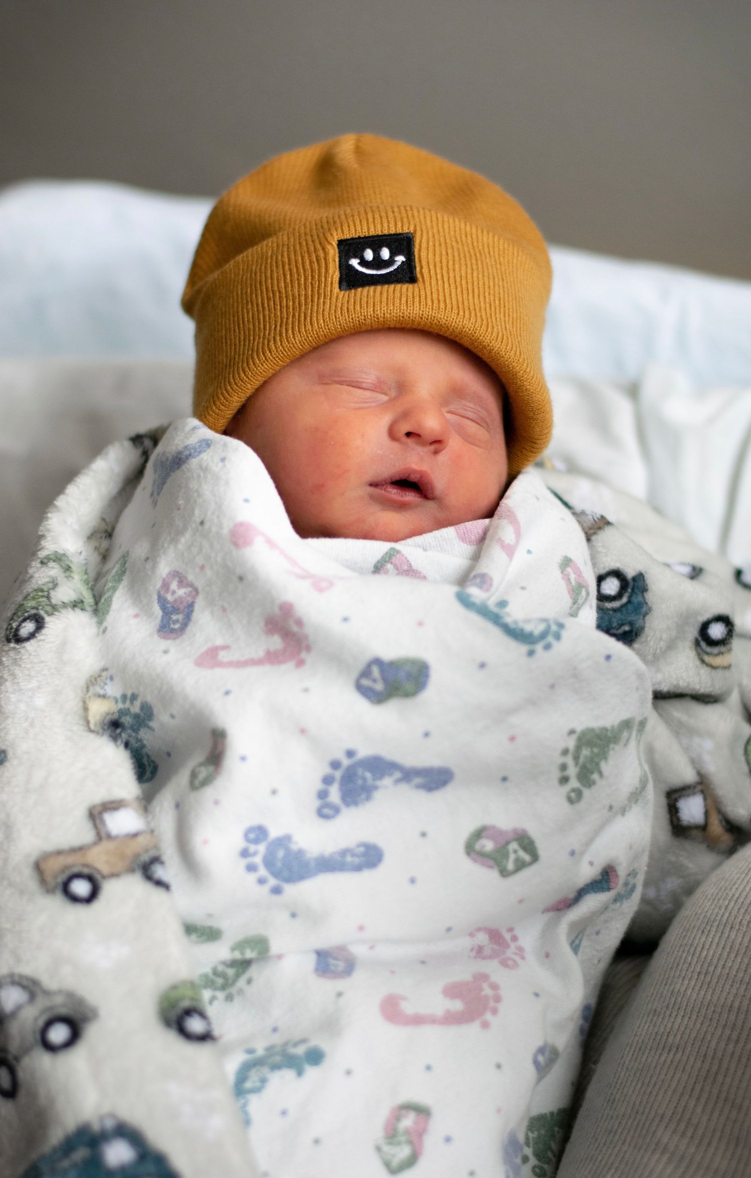 A sleeping newborn baby wearing a mustard yellow beanie is swaddled in a white blanket with colorful vehicle print pattern.