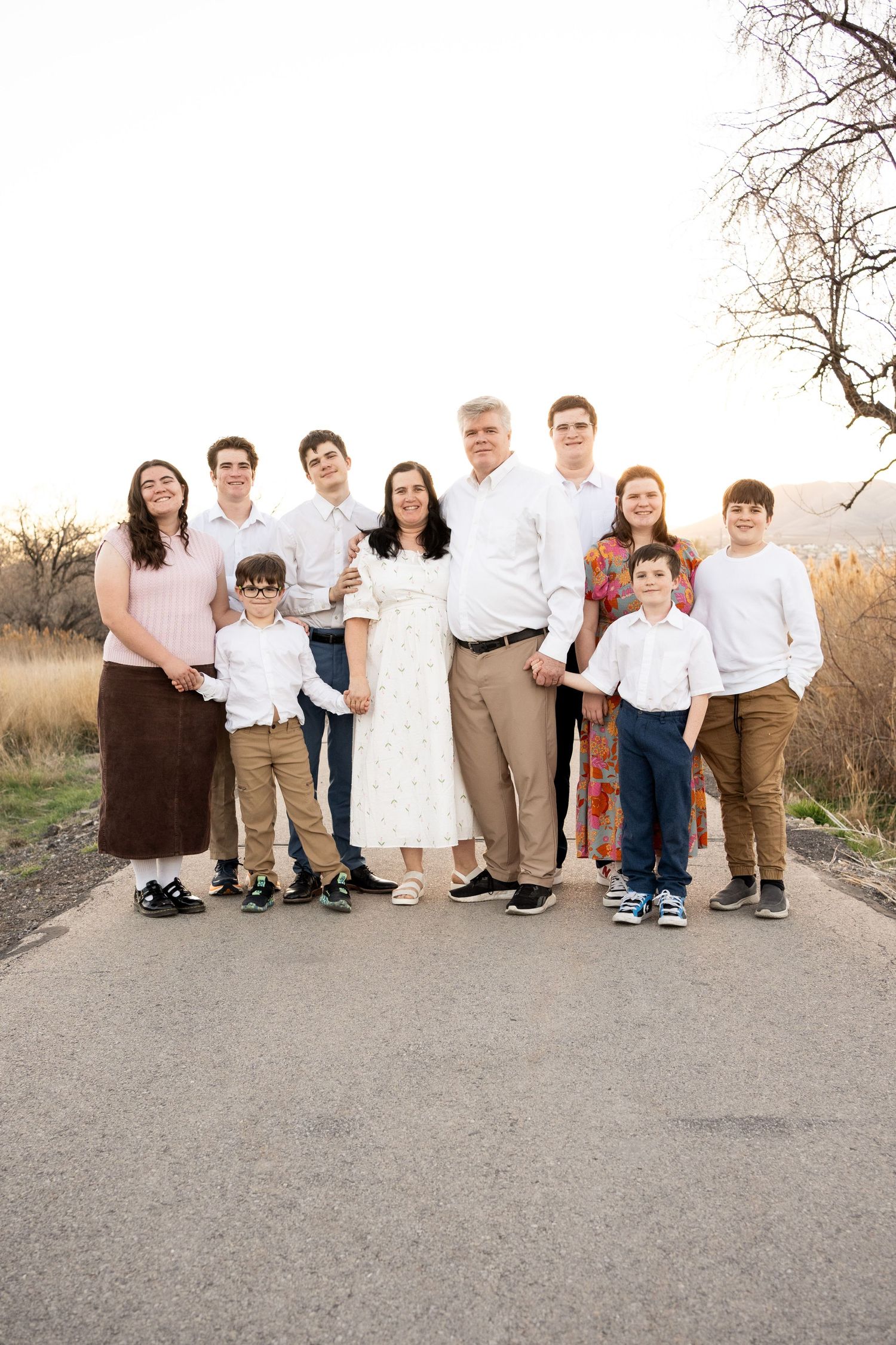 Large family of ten posing together on a path outdoors during a bright day in coordinated neutral outfits.