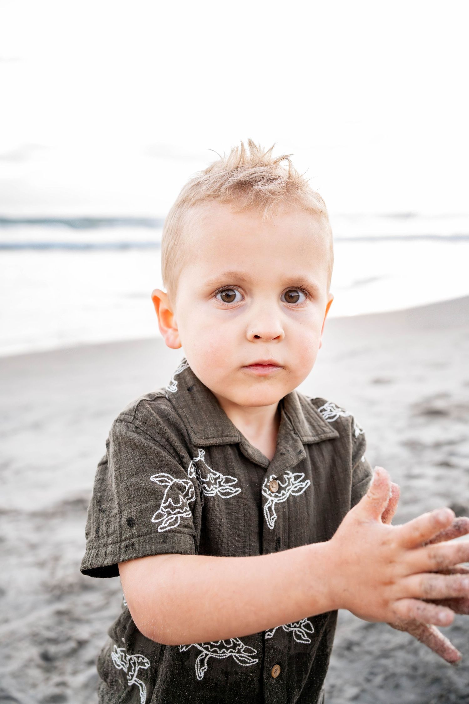A young blonde toddler boy in a dark floral shirt stands on a sandy beach with ocean waves in the background.