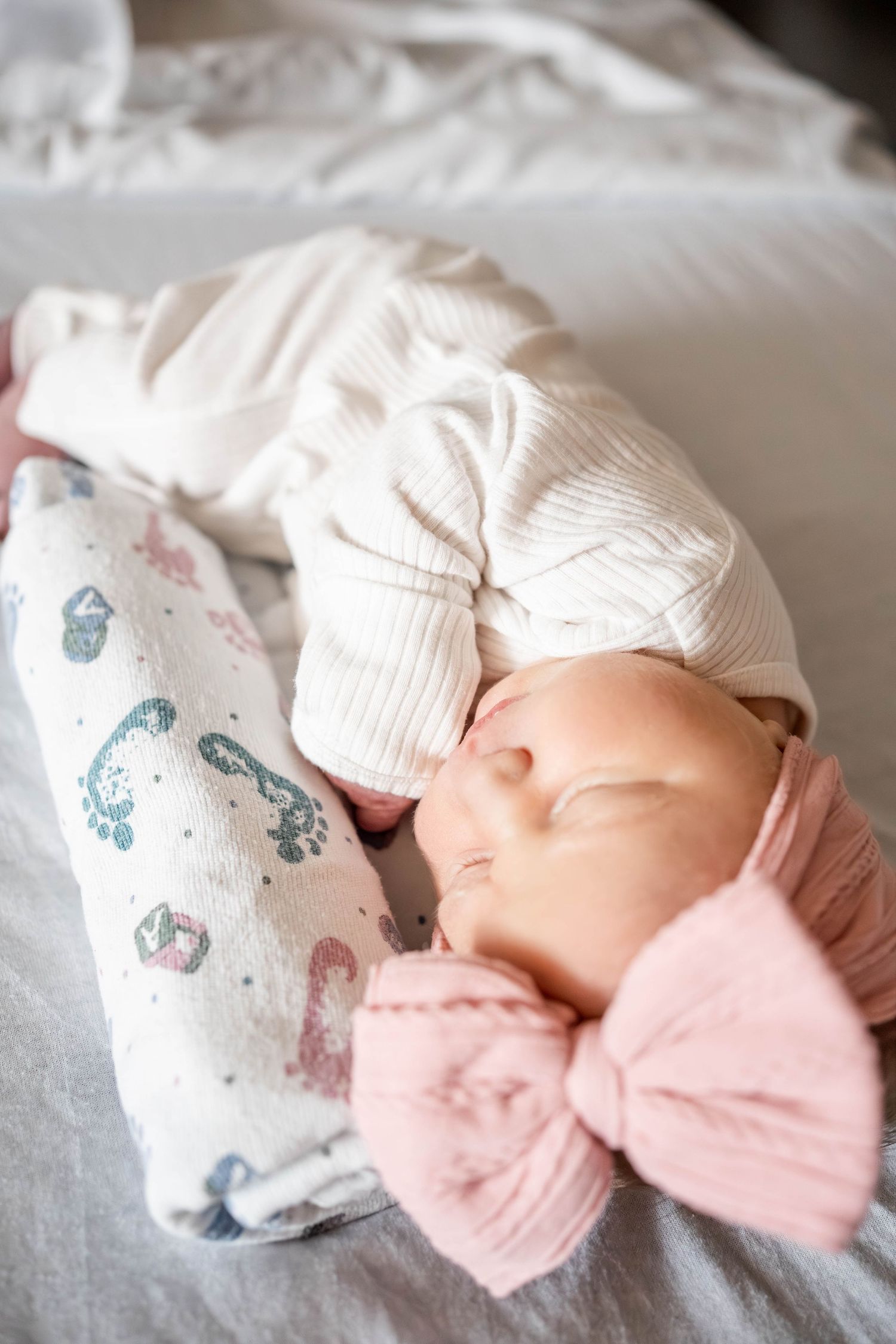 Newborn baby sleeping peacefully in white ribbed outfit and pink bow headband on a soft gray bed.