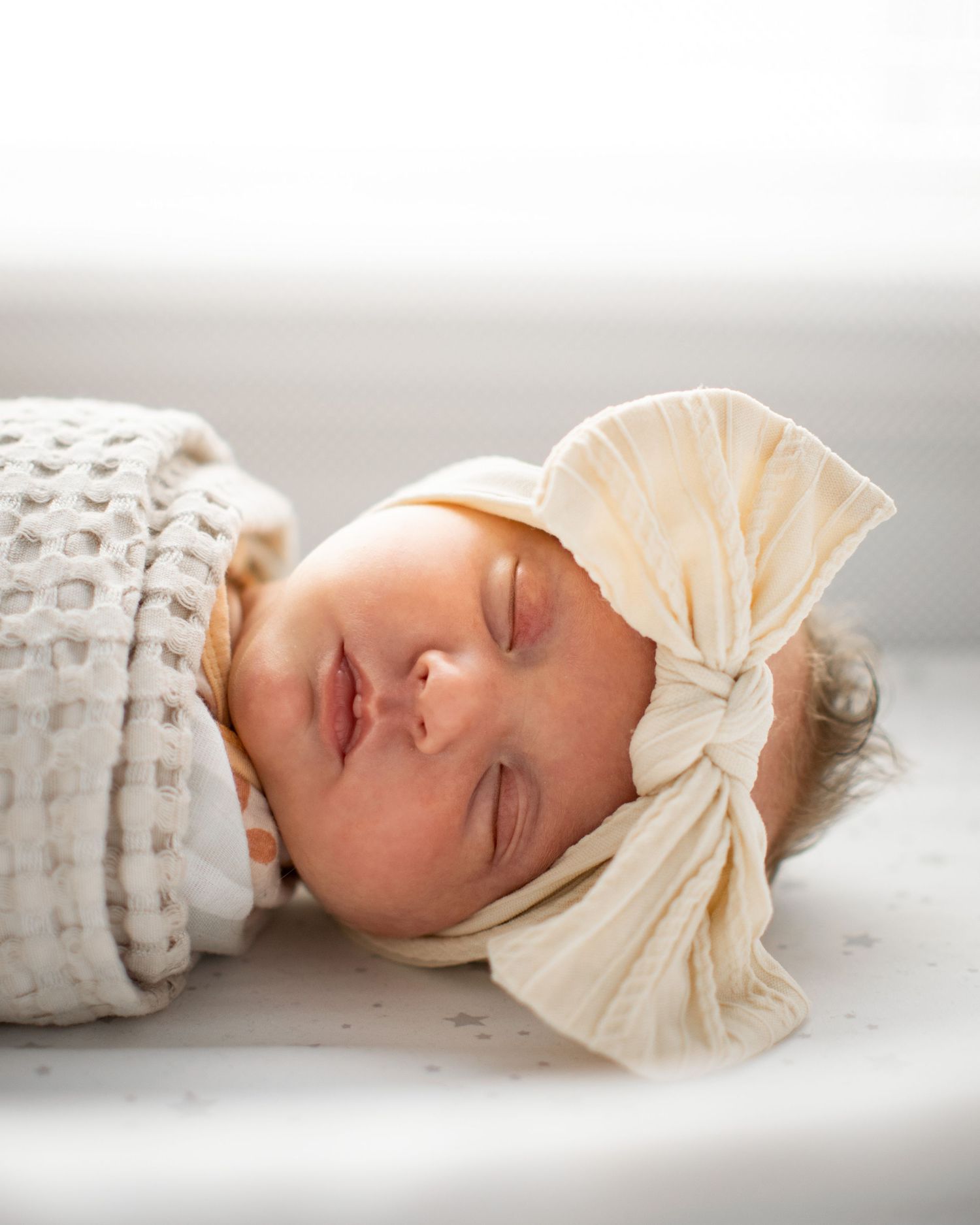 Baby wearing cream bow headband lying peacefully on white textured surface.