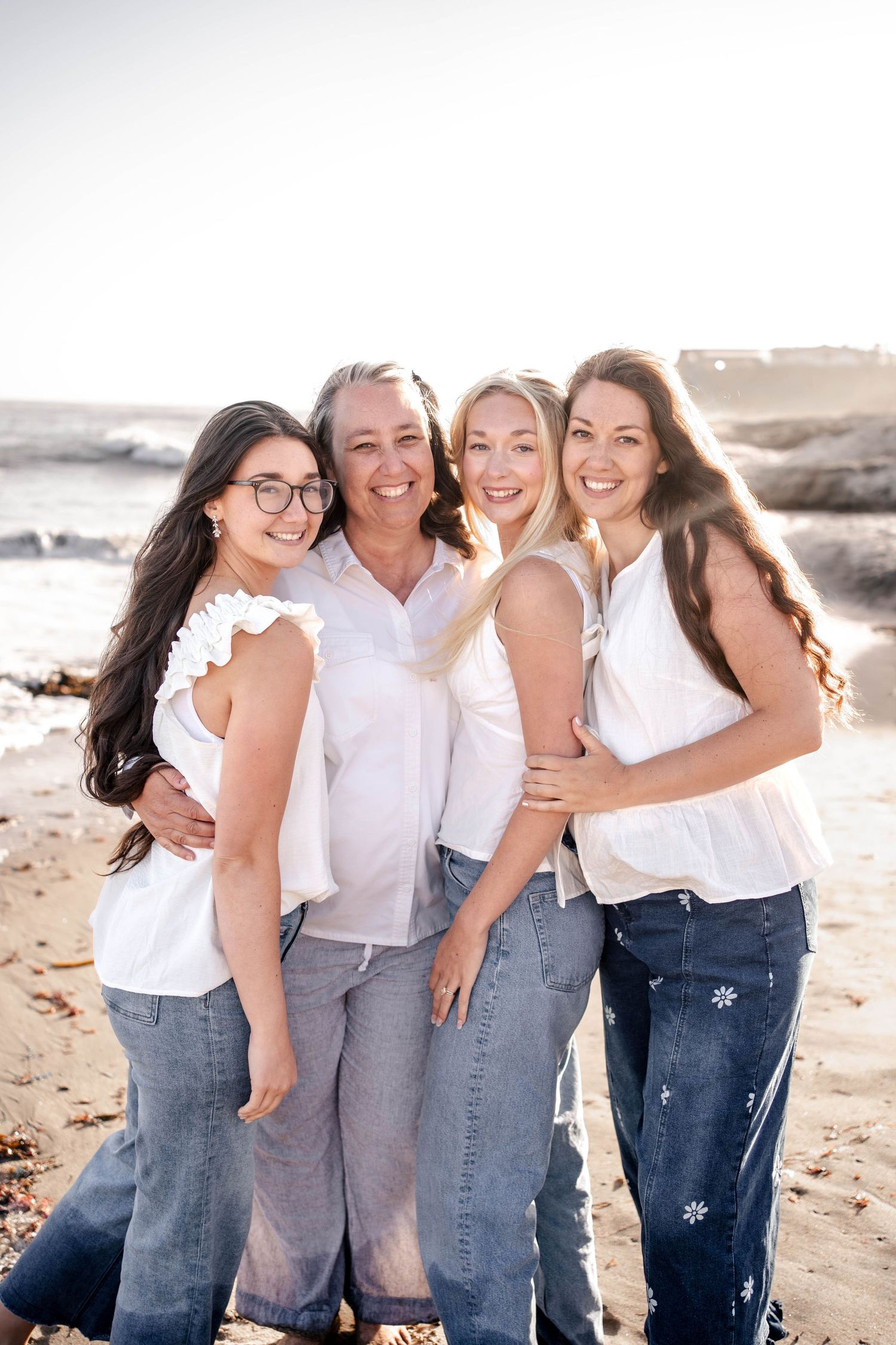 Four women in white tops and jeans smile together on a sunny beach, posing for a family portrait.