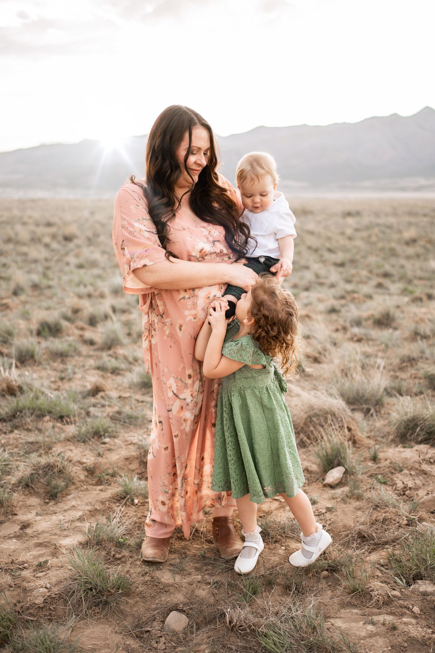 Mother in floral dress holds baby while toddler in green dress reaches up during golden hour desert photoshoot.