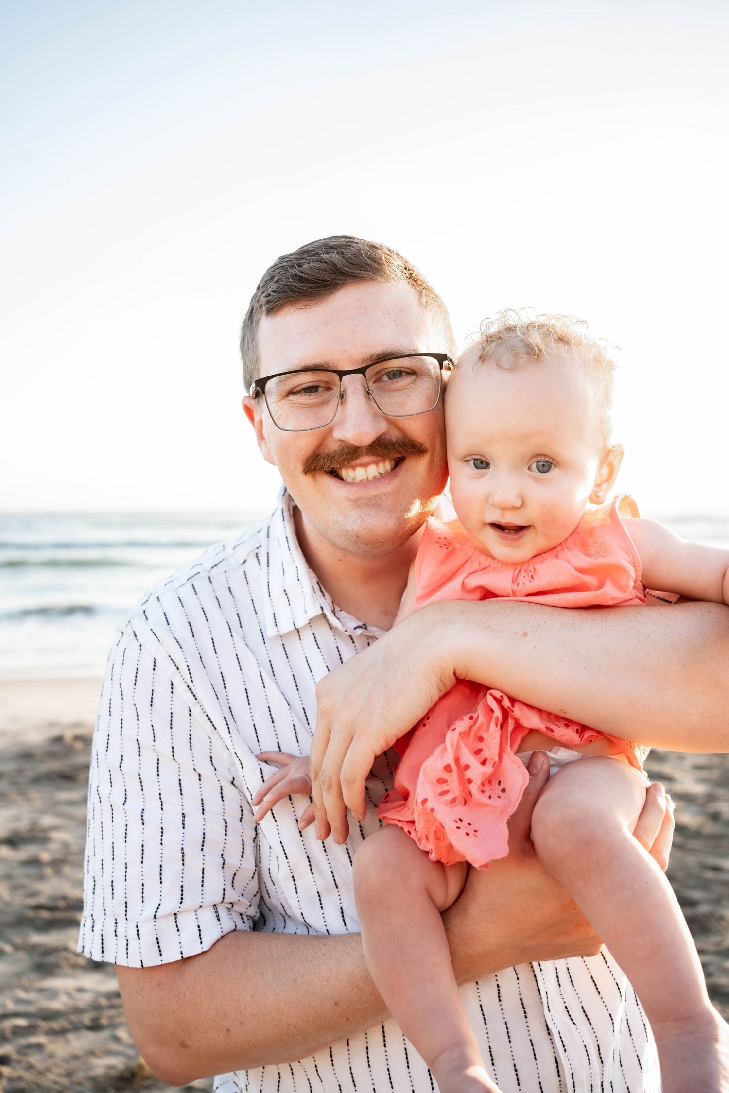Smiling man with glasses and mustache holds a baby girl in pink dress on a sunny beach.