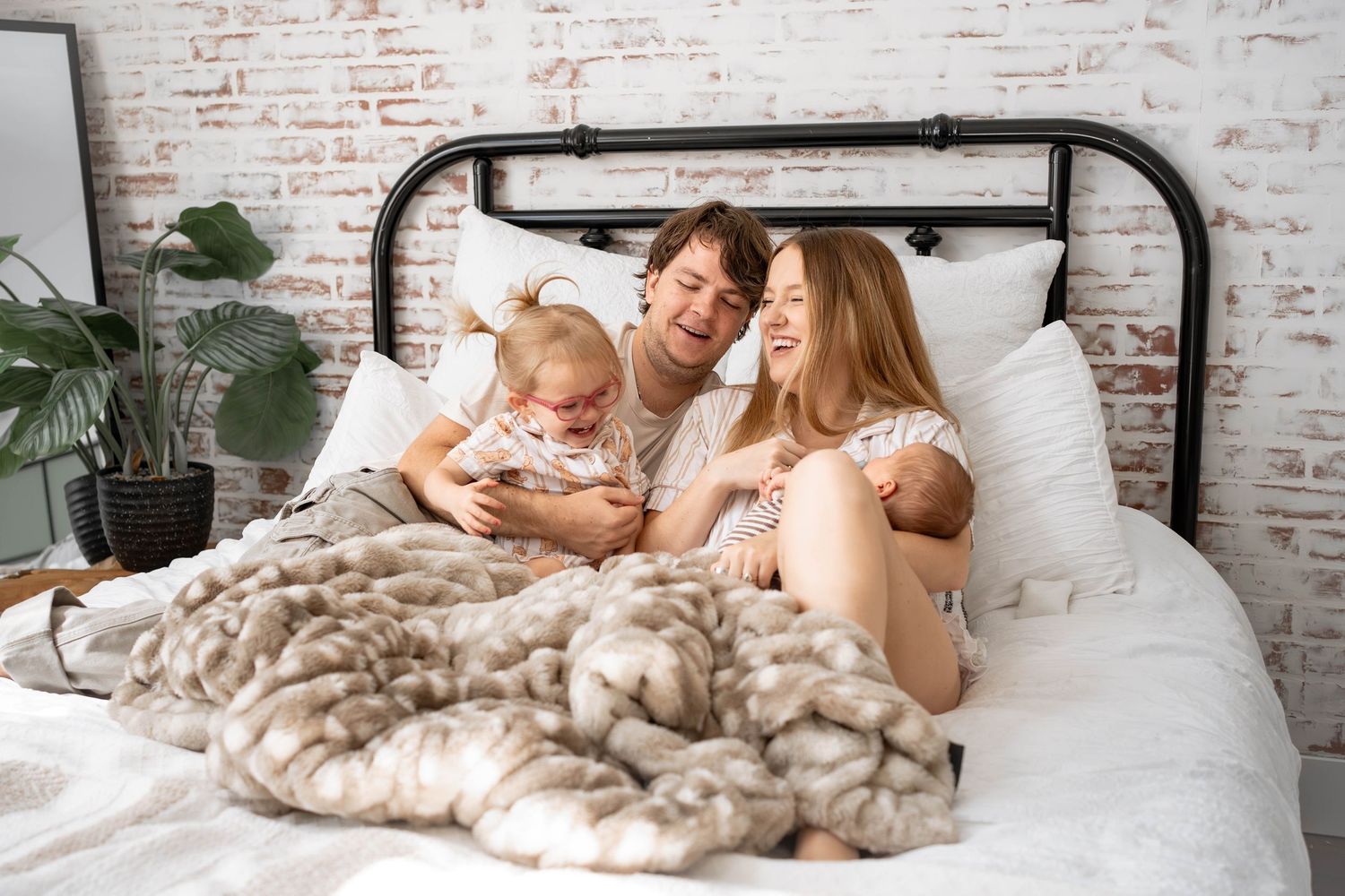 Smiling family of four relaxing together on a white bed with a black metal frame, cozy fur blanket, and exposed brick wall.