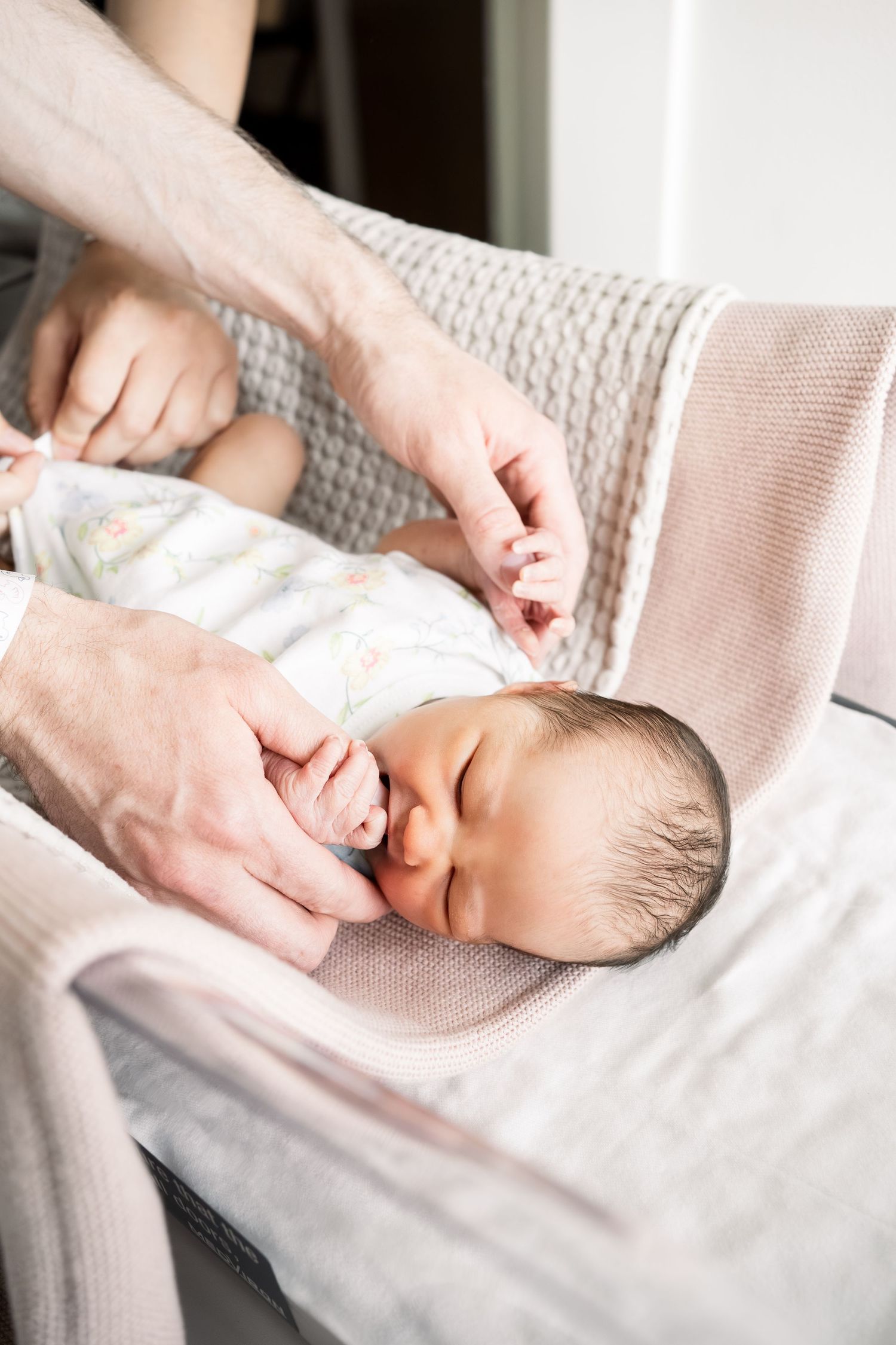 Newborn baby sleeping peacefully in a bouncer while an adult gently fastens the safety harness.