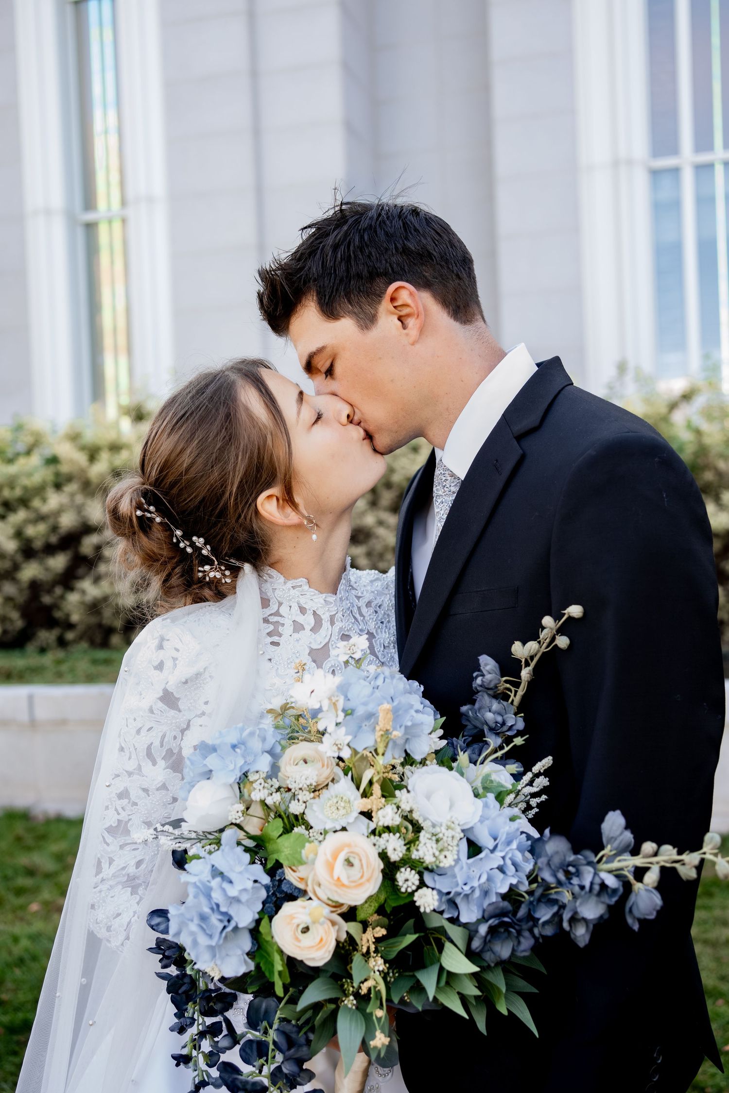 Bride in a lace gown and veil kisses her groom in a dark suit while holding a blue and cream floral bouquet outside a temple.