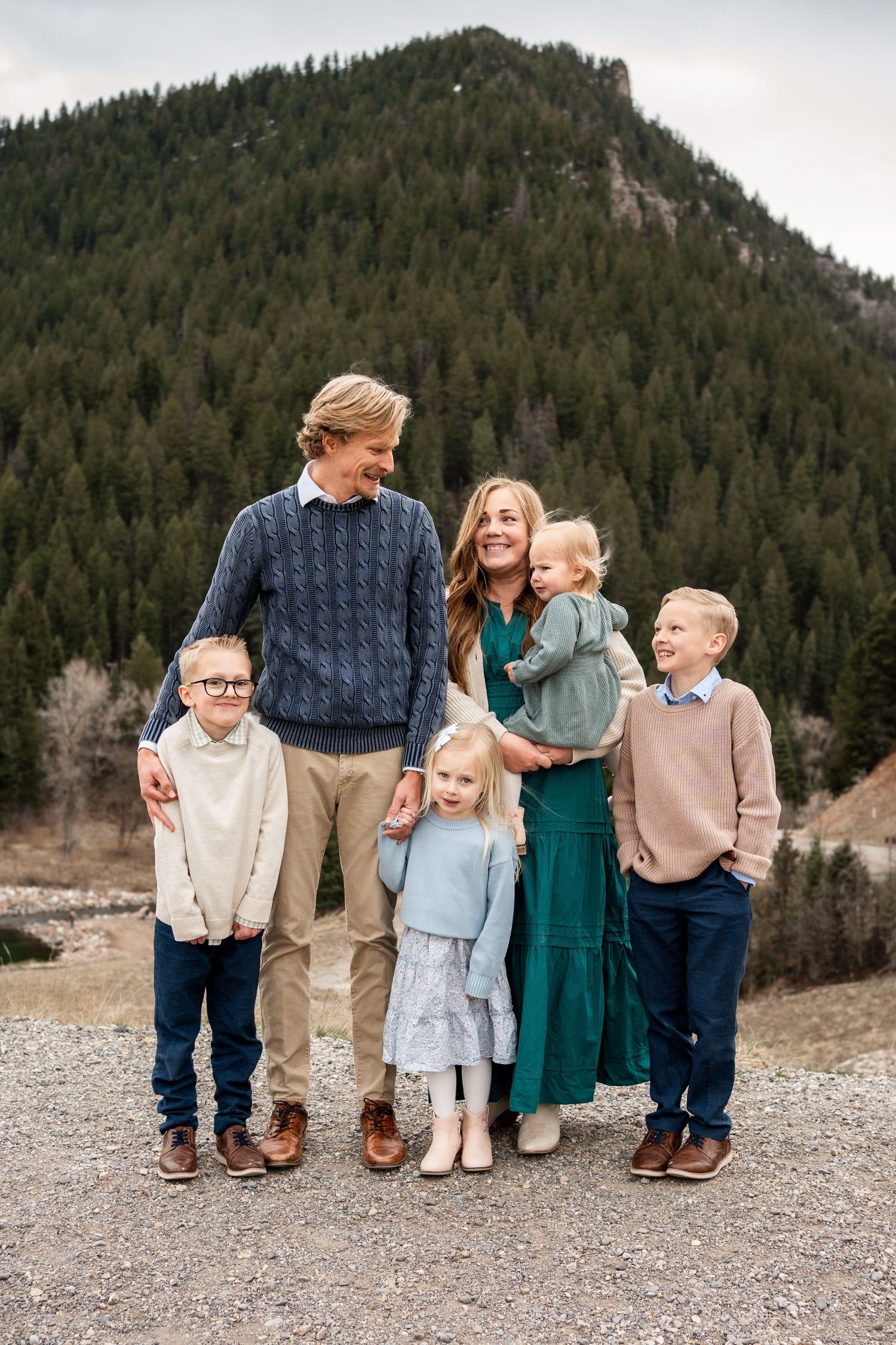 A family stands together against a scenic mountain backdrop on a rocky shore wearing coordinated casual outfits.