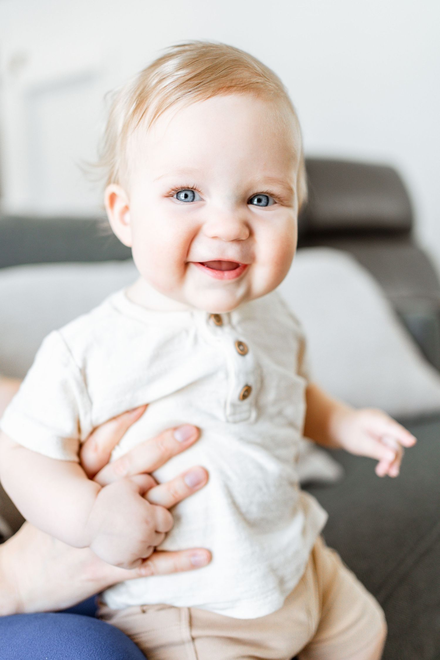 A smiling baby wearing a white button-up shirt and beige pants sits supported on a gray couch.