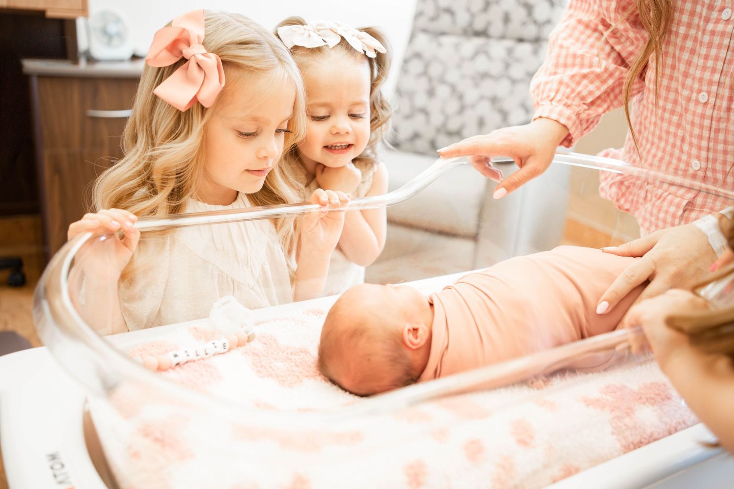 Two young girls with bows admire a swaddled newborn baby in a hospital bassinet.