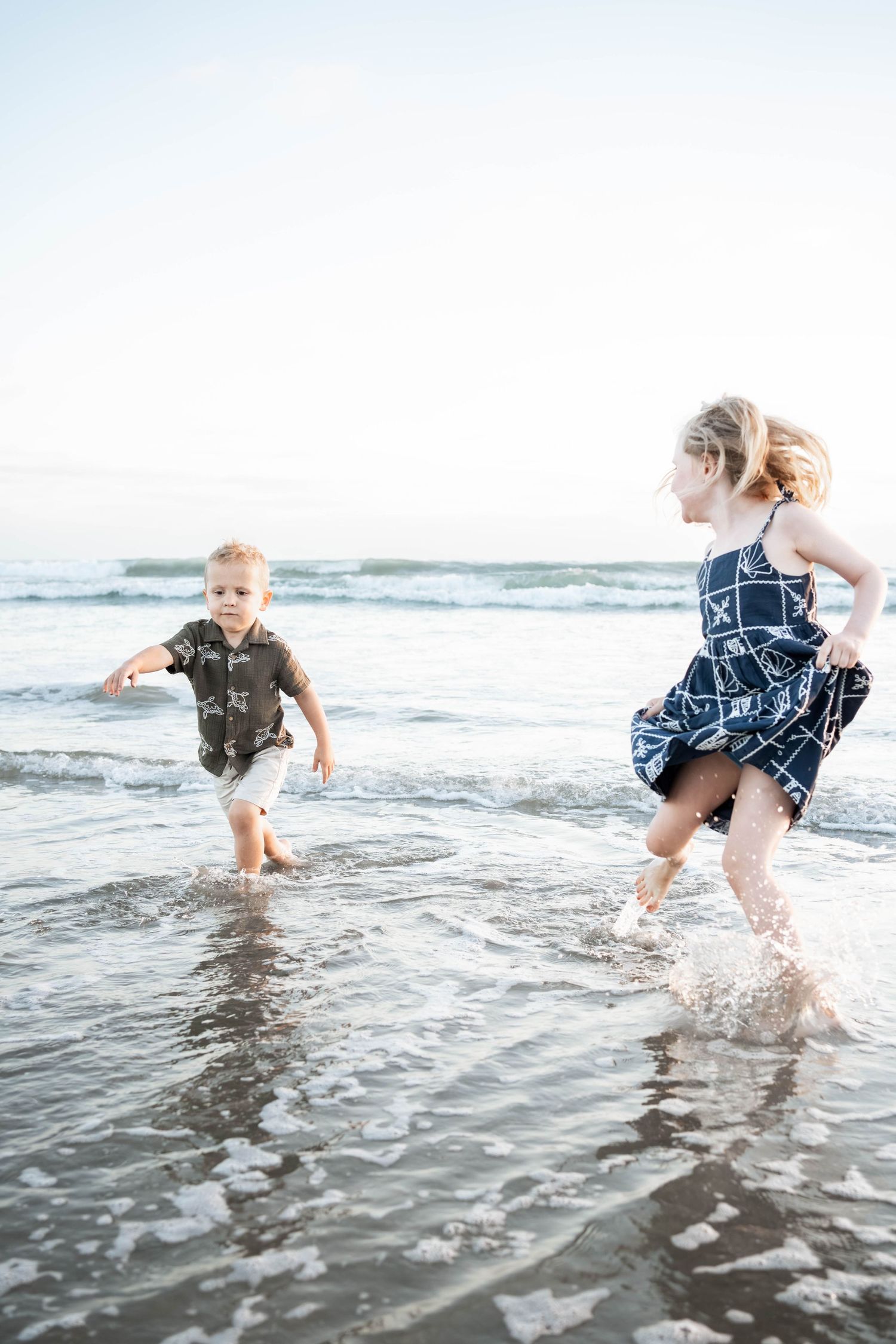 Two young children splashing and running in ocean waves on a sandy beach during a sunny day.