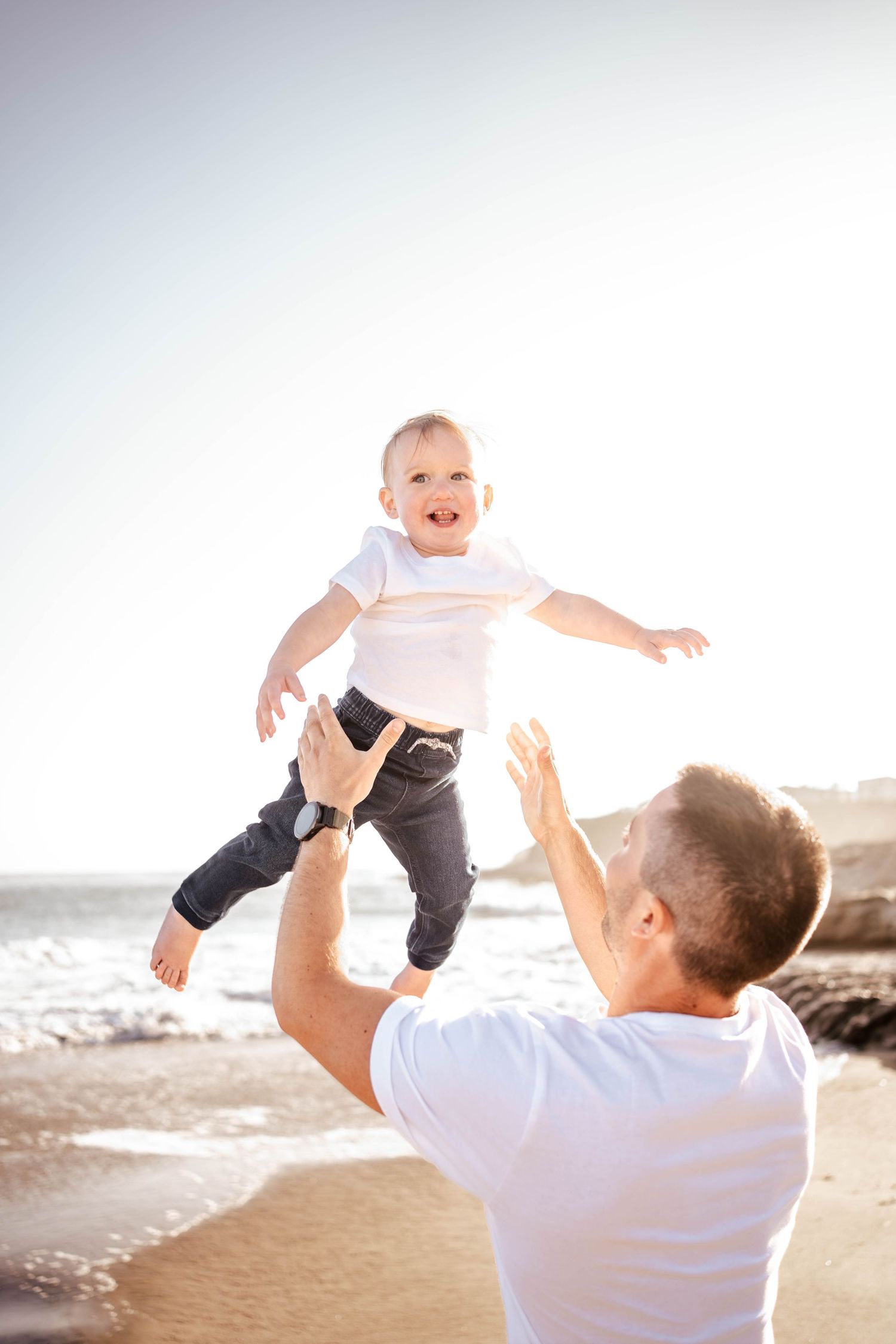 Father joyfully tosses smiling baby in the air on a sunny beach, with ocean waves in the background.