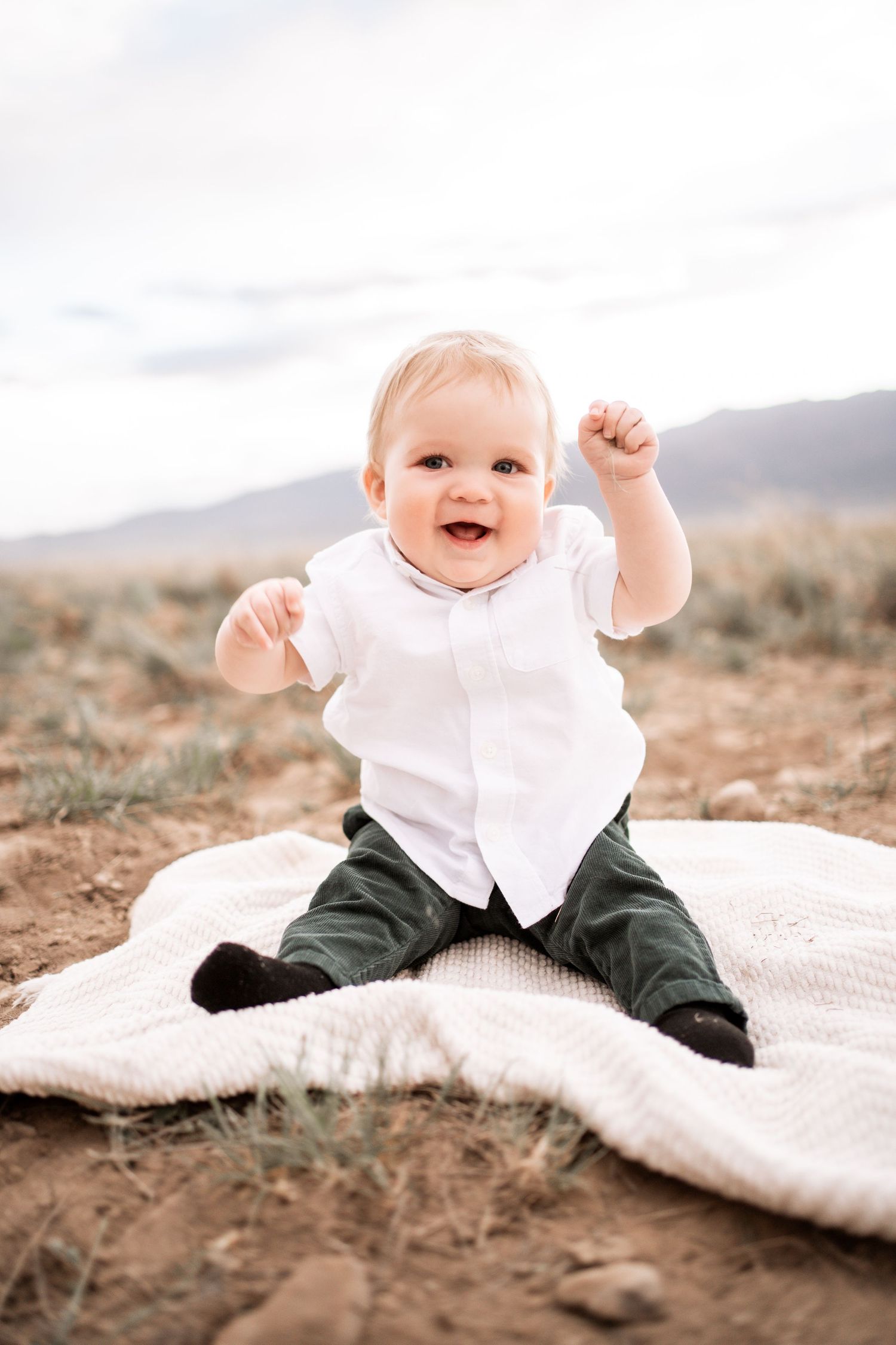 A happy baby in a white shirt and green pants sits on a blanket outdoors with arms raised joyfully.