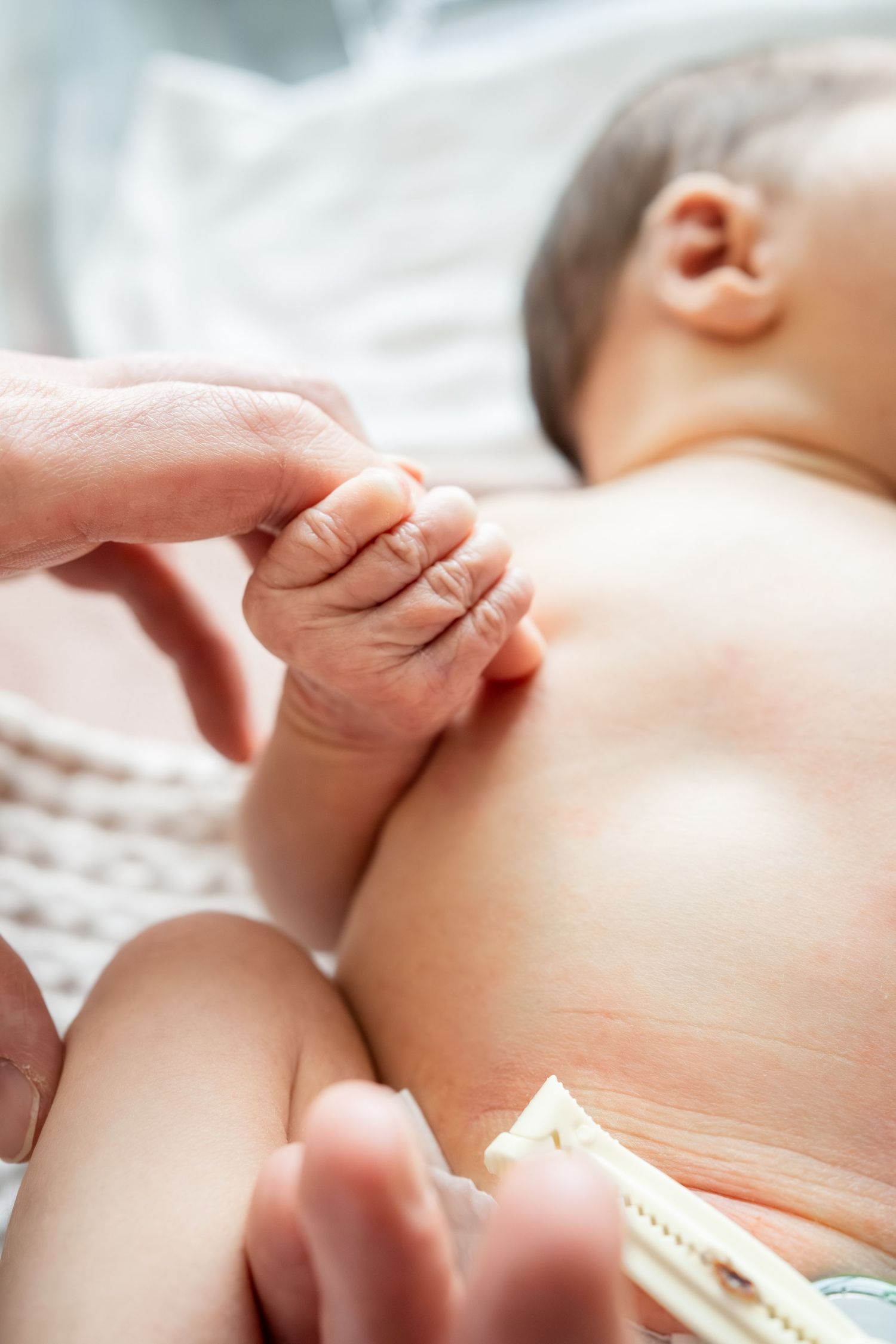 Newborn baby grasping adult finger, lying on white blanket with umbilical cord clamp visible.