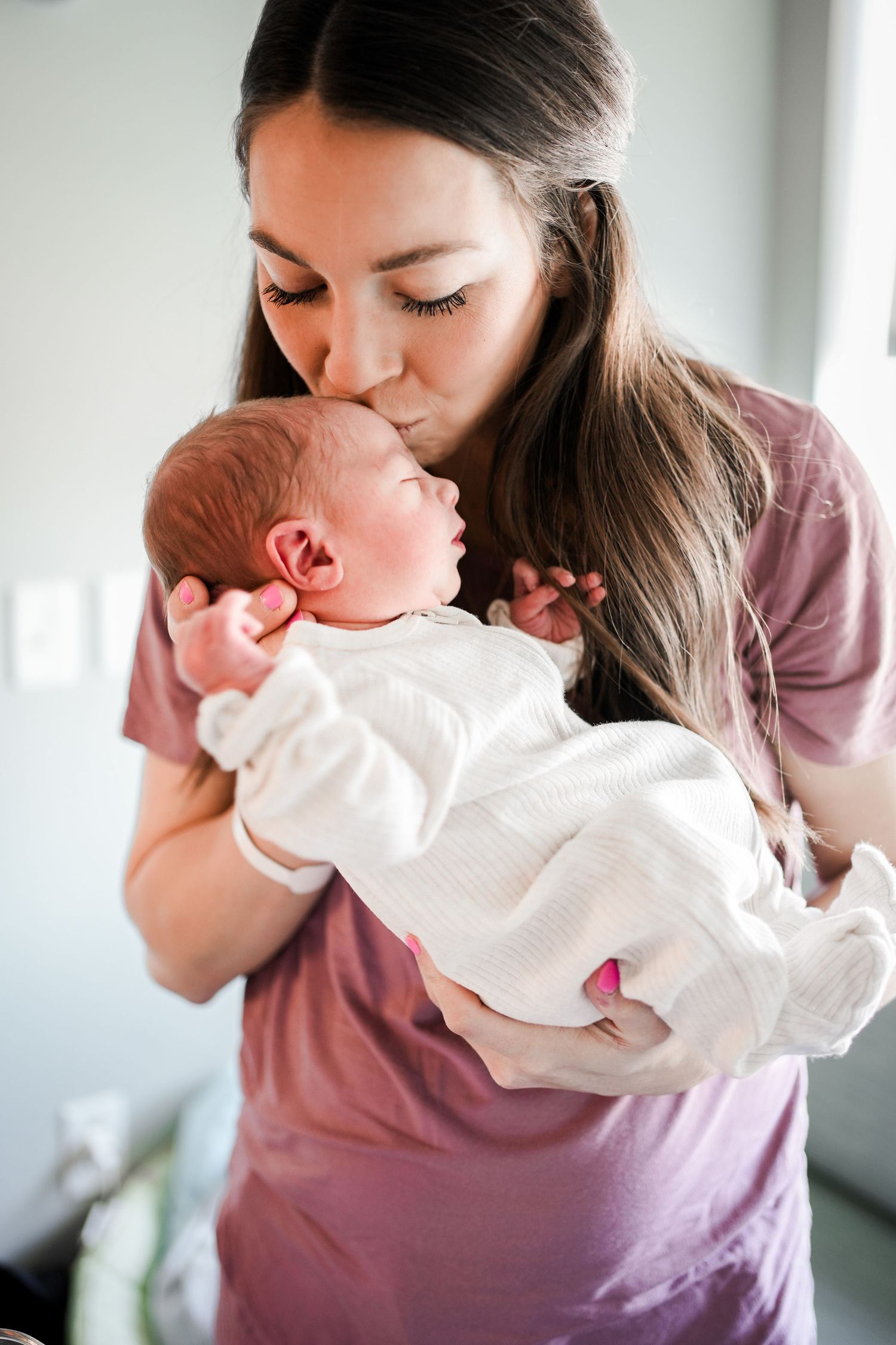 A tender moment between mother and baby as she gently kisses the infant while holding them in a mauve colored shirt.