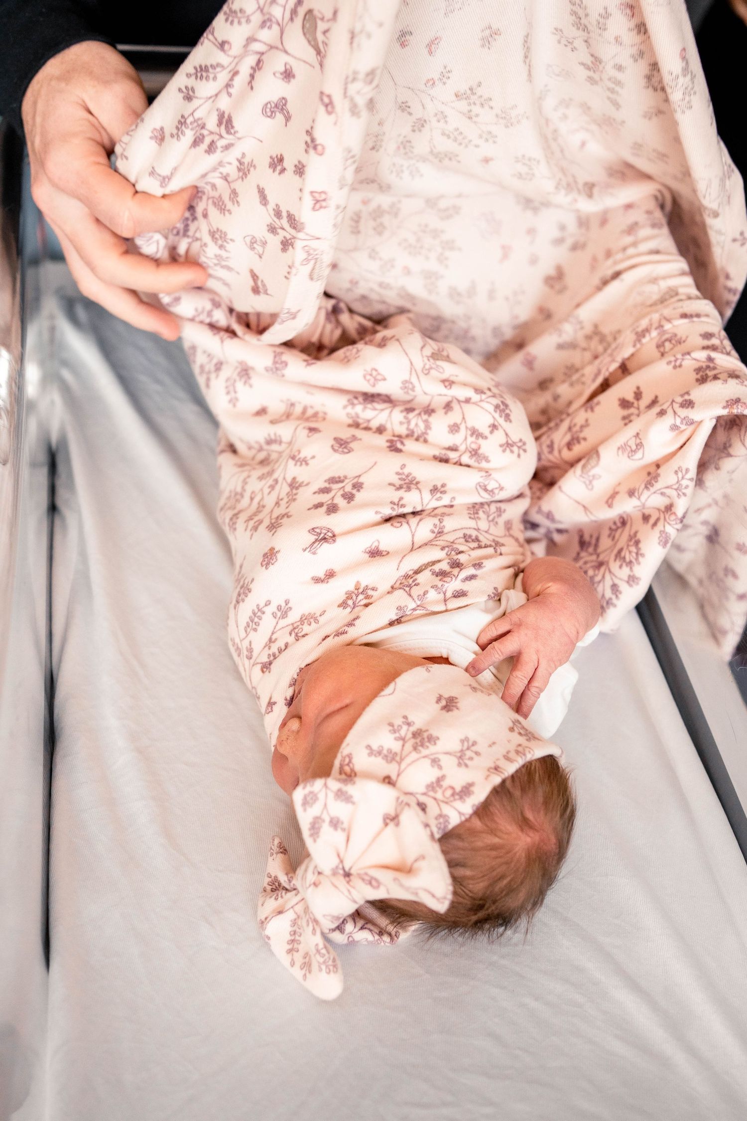 Newborn baby lying in hospital bassinet wrapped in floral pink swaddle blanket with matching bow headband.