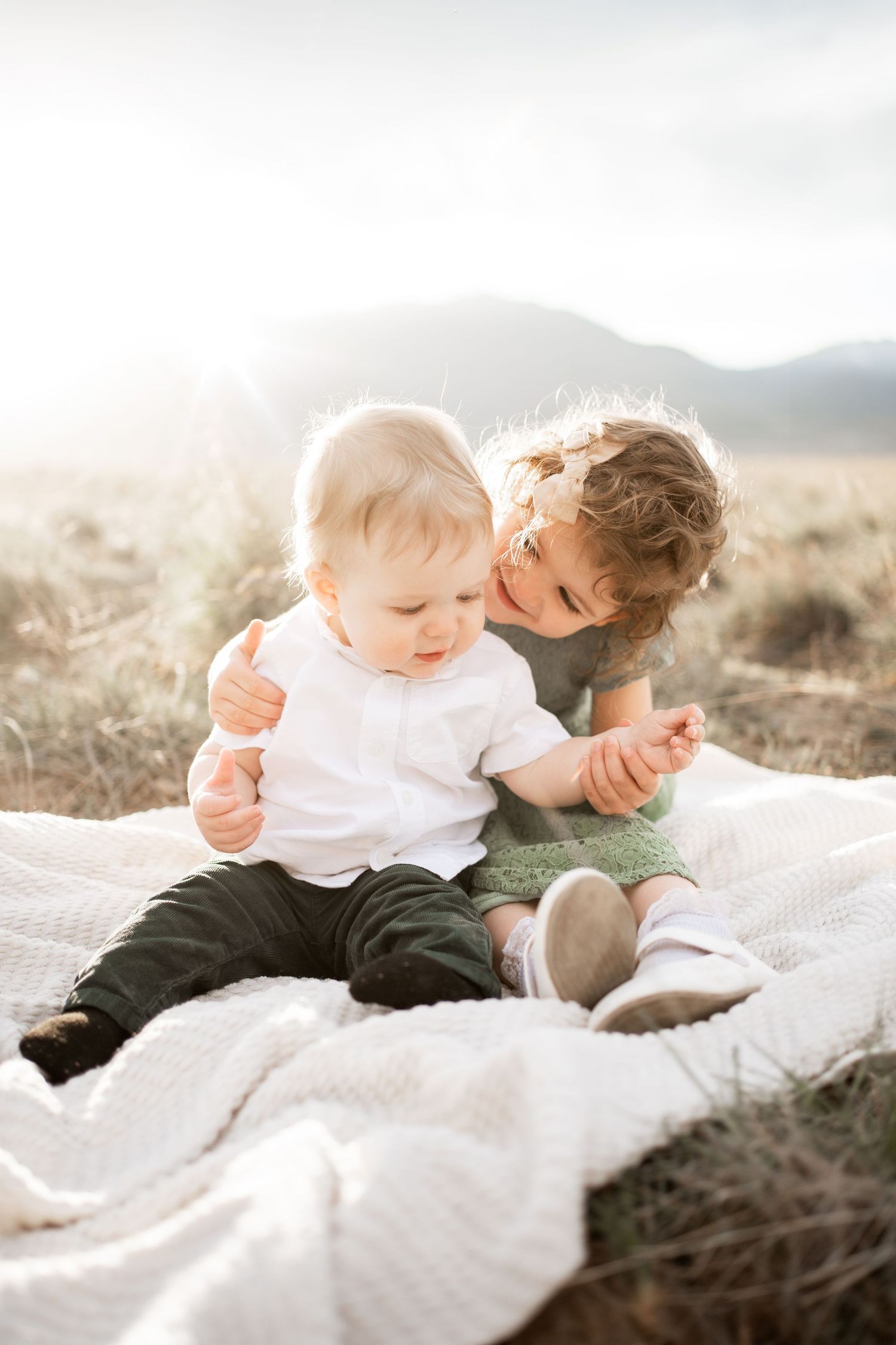 Two young siblings sit on a blanket in a sunlit field, with the older child lovingly embracing the baby.