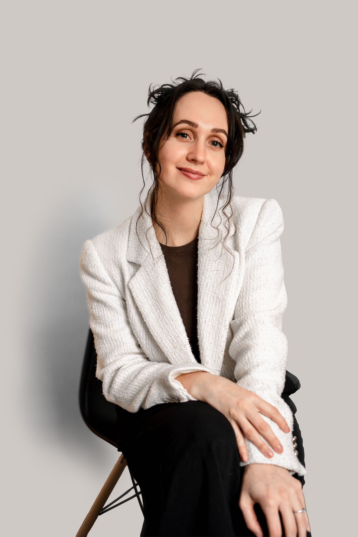 A confident young woman in a white blazer and black pants sits gracefully on a chair against a light gray background.