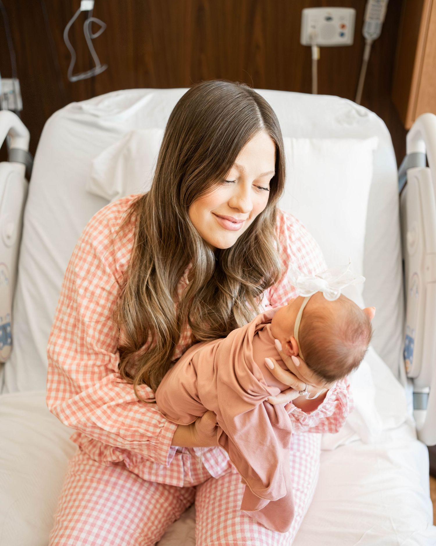A smiling new mother in pink gingham pajamas cradles her newborn baby in a hospital room.