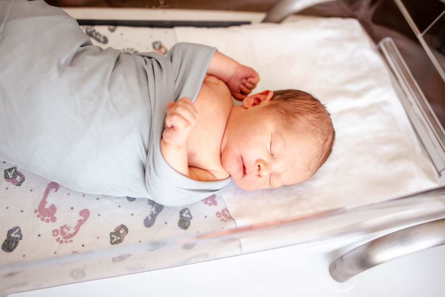 Newborn baby swaddled in gray blanket sleeping peacefully in hospital bassinet.