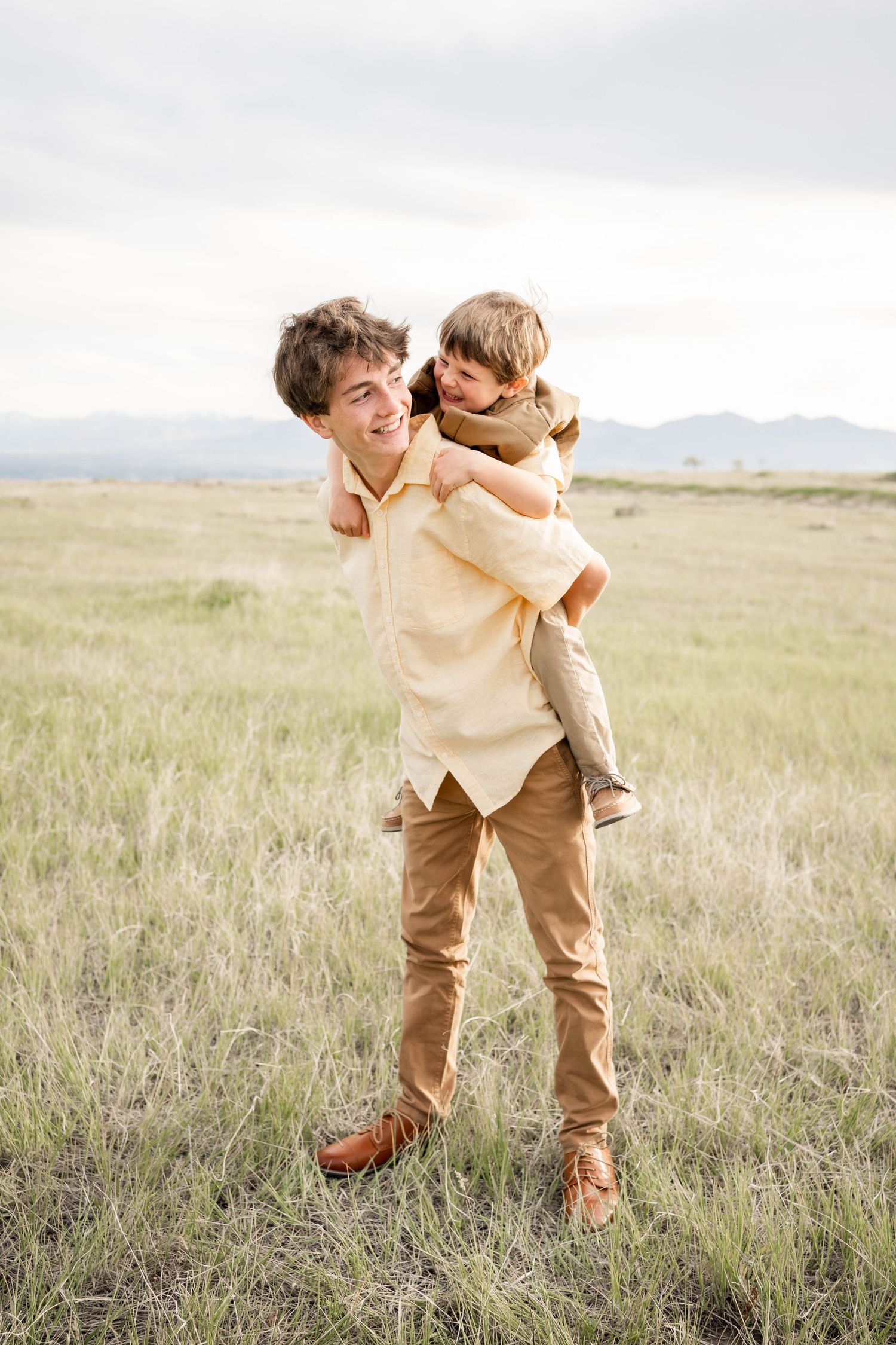 A young man gives a child a piggyback ride in a grassy field with mountains in the background.
