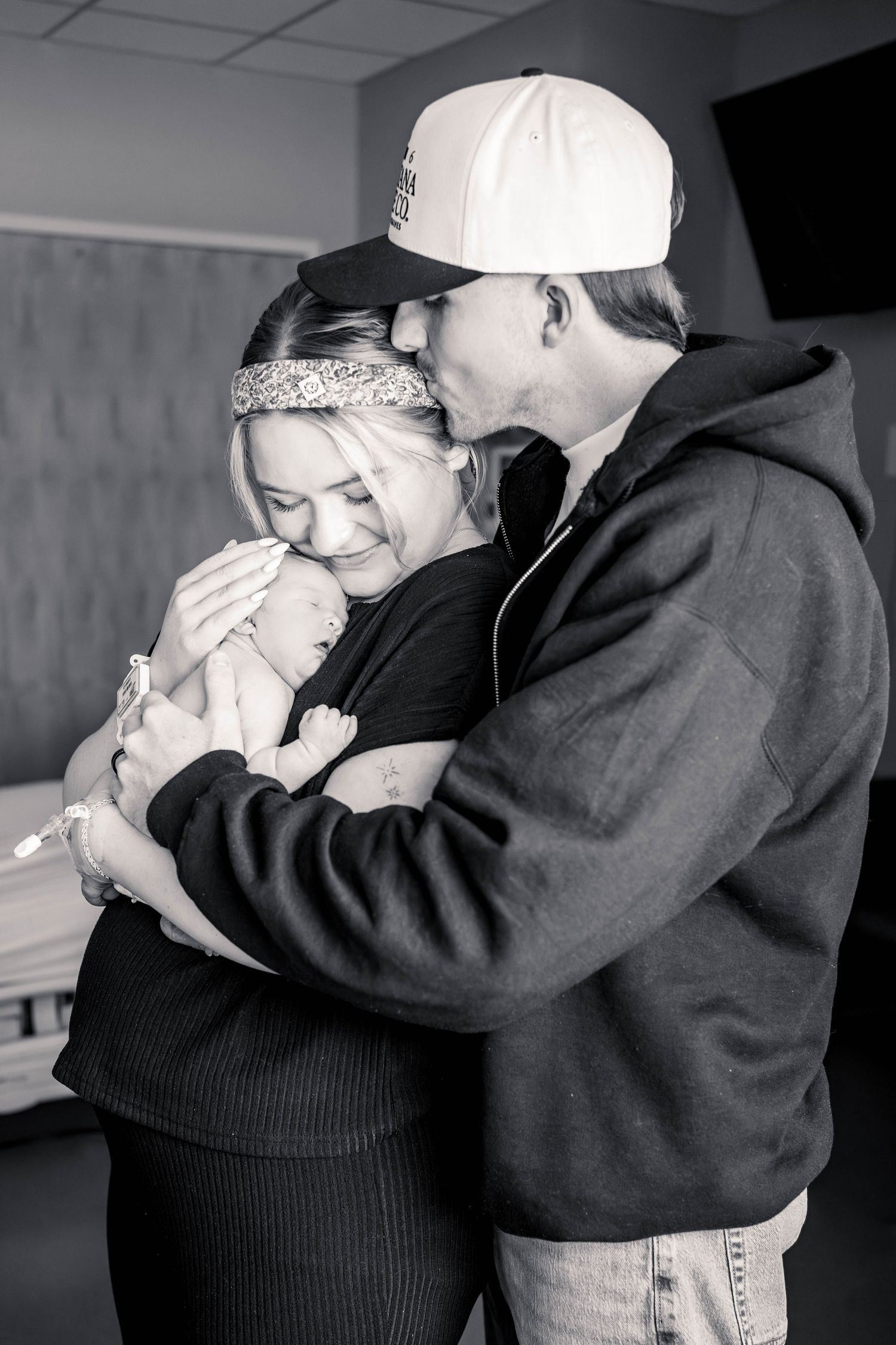 Black and white photo of new parents holding their newborn baby in a hospital room, father kissing mothers head.