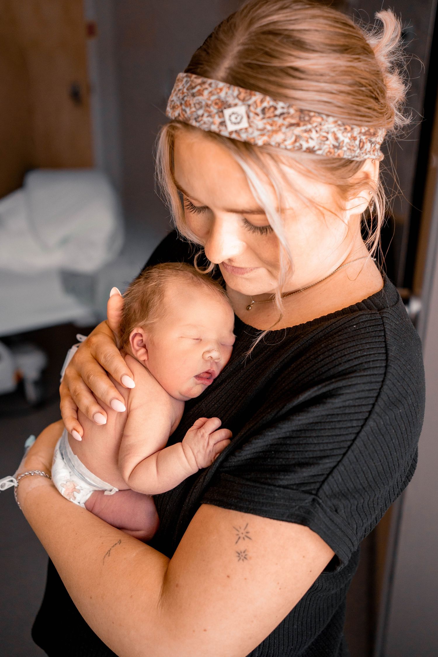 Young mother with floral headband tenderly cradling her sleeping newborn baby in a warm indoor setting.