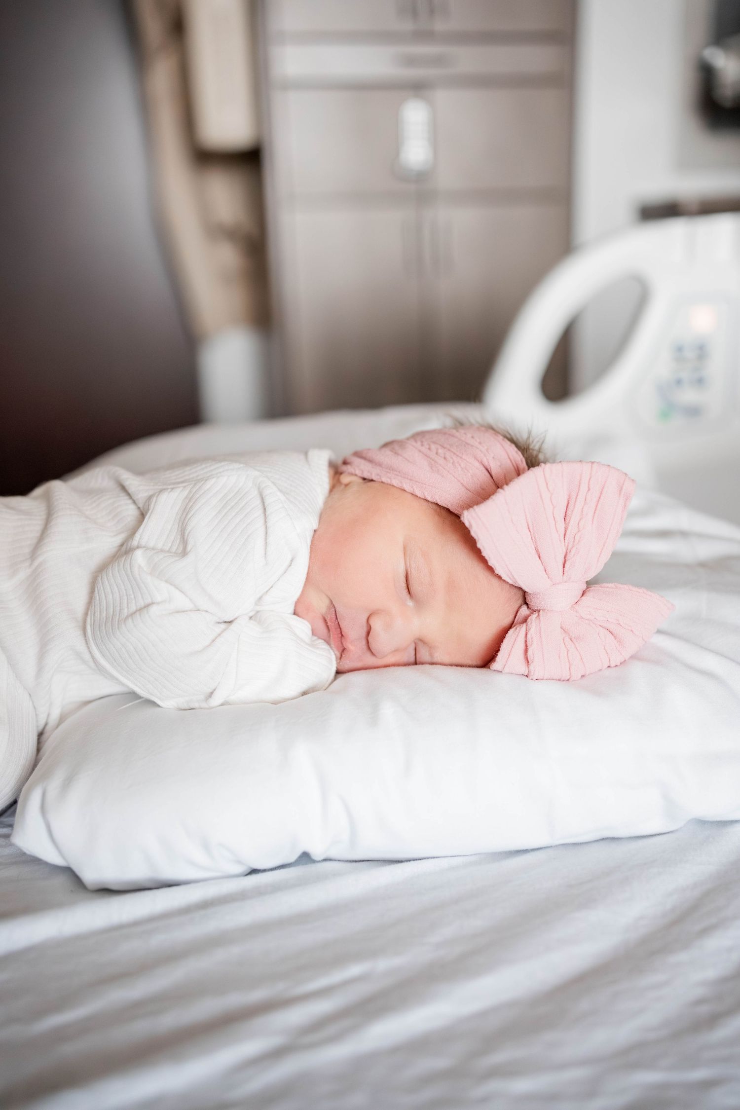 Newborn baby girl sleeping peacefully on white pillow wearing pink bow headband and white swaddle in hospital room.