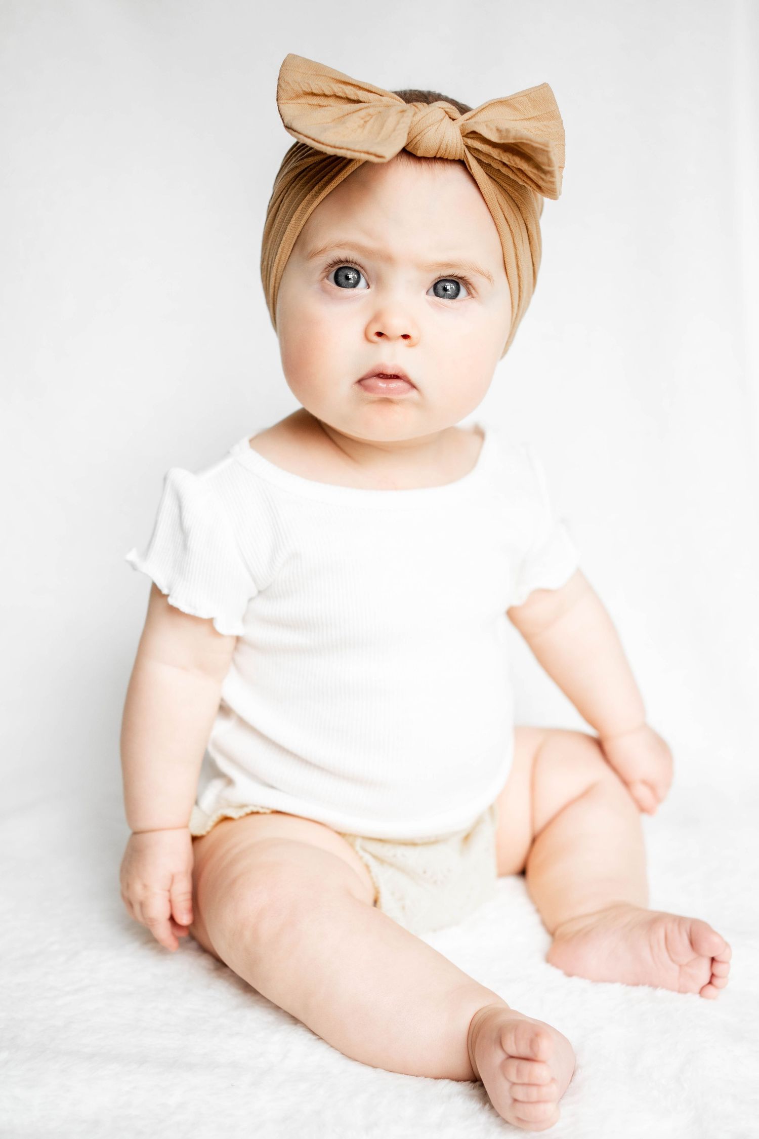 Baby girl in a tan knotted bow headband and white ruffle-sleeve bodysuit sits barefoot on a soft white blanket.