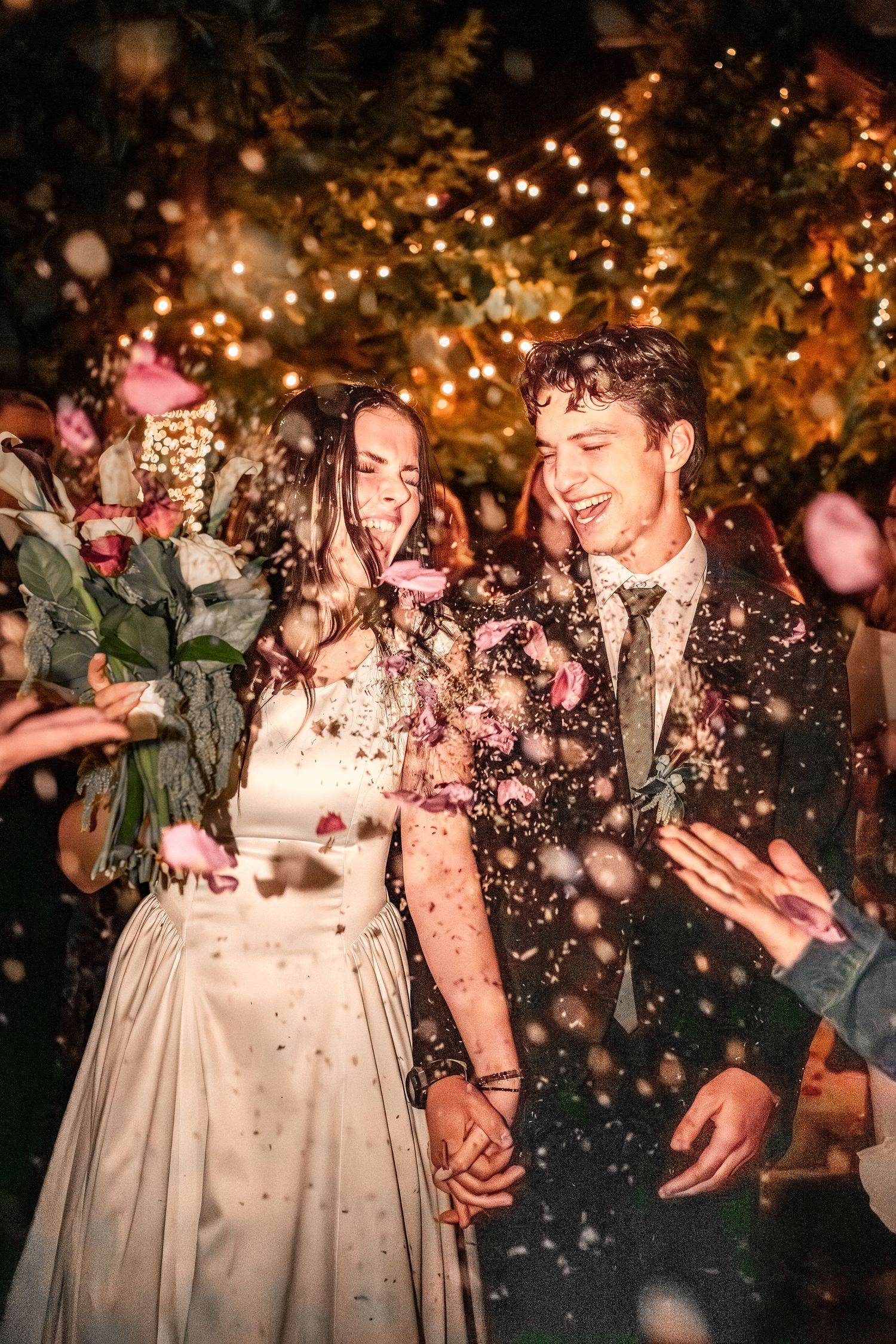 Bride and groom laugh while holding hands as rose petals fly around them at a nighttime wedding exit under glowing string lights.