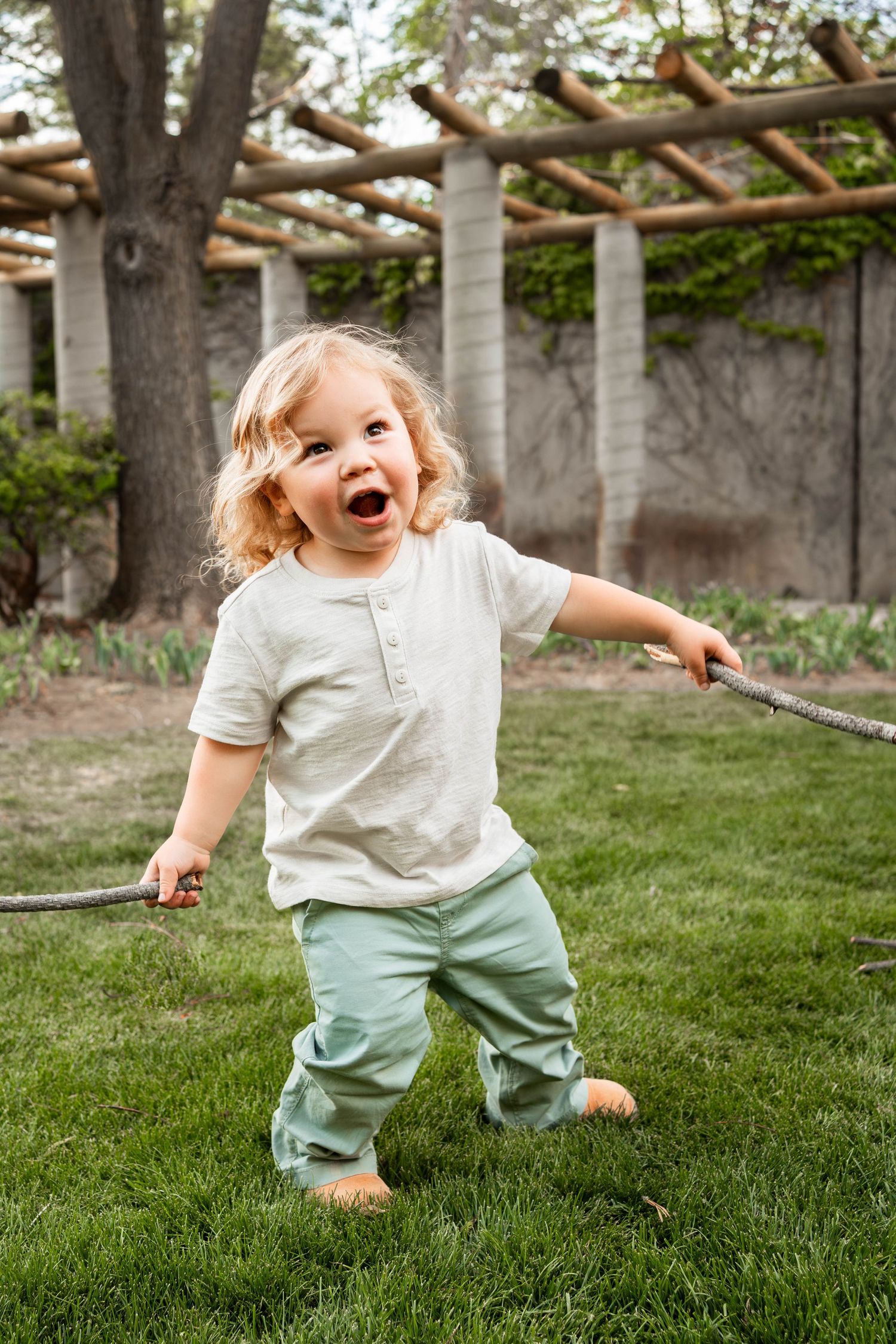 A smiling curly-haired toddler in a white shirt and sage green pants holds a long stick while playing on a grassy lawn.