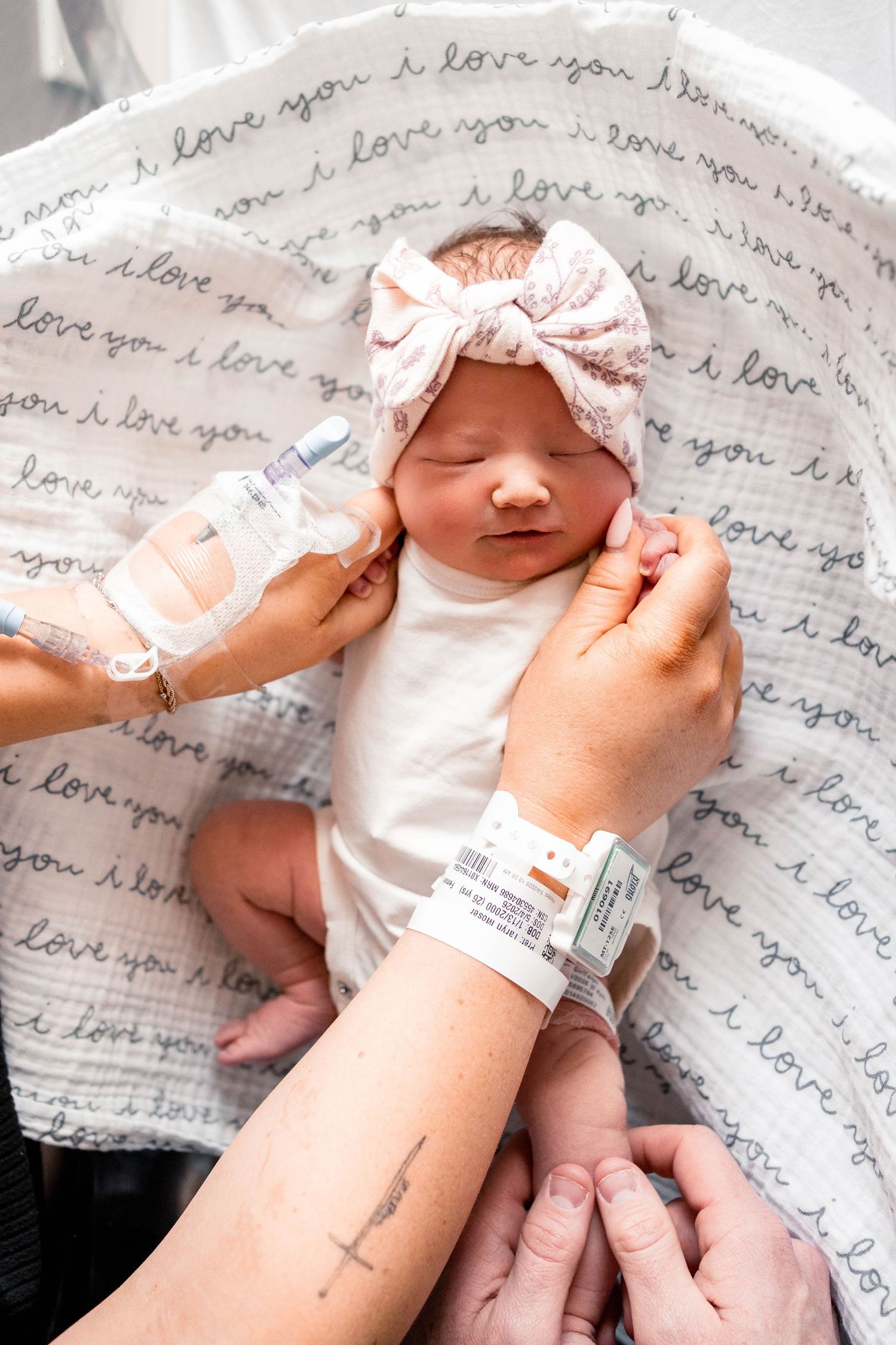 Newborn baby girl in white onesie and pink bow headband lying on love-print blanket in hospital.