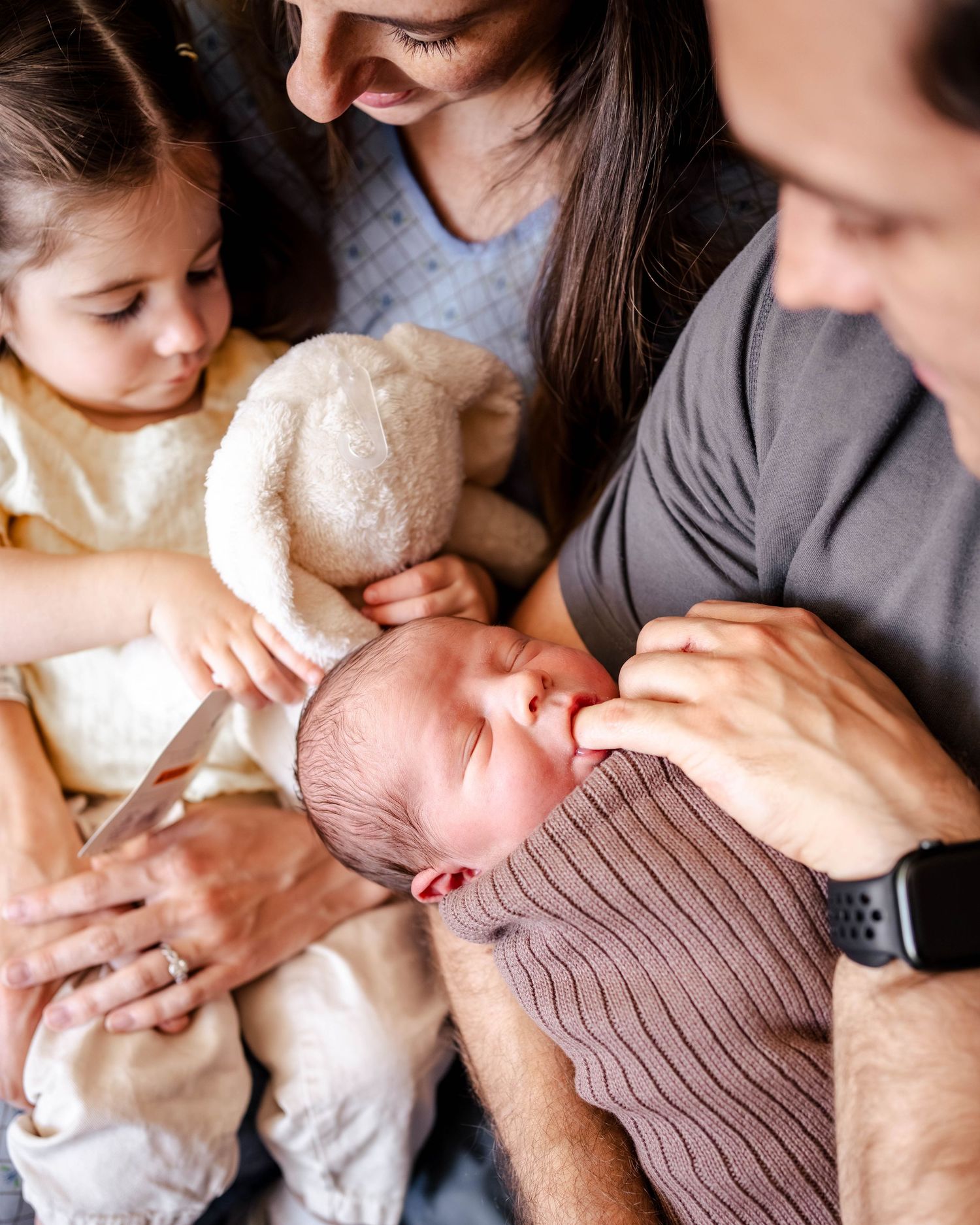 Family gathered around a swaddled newborn baby, with a toddler holding a stuffed animal nearby.