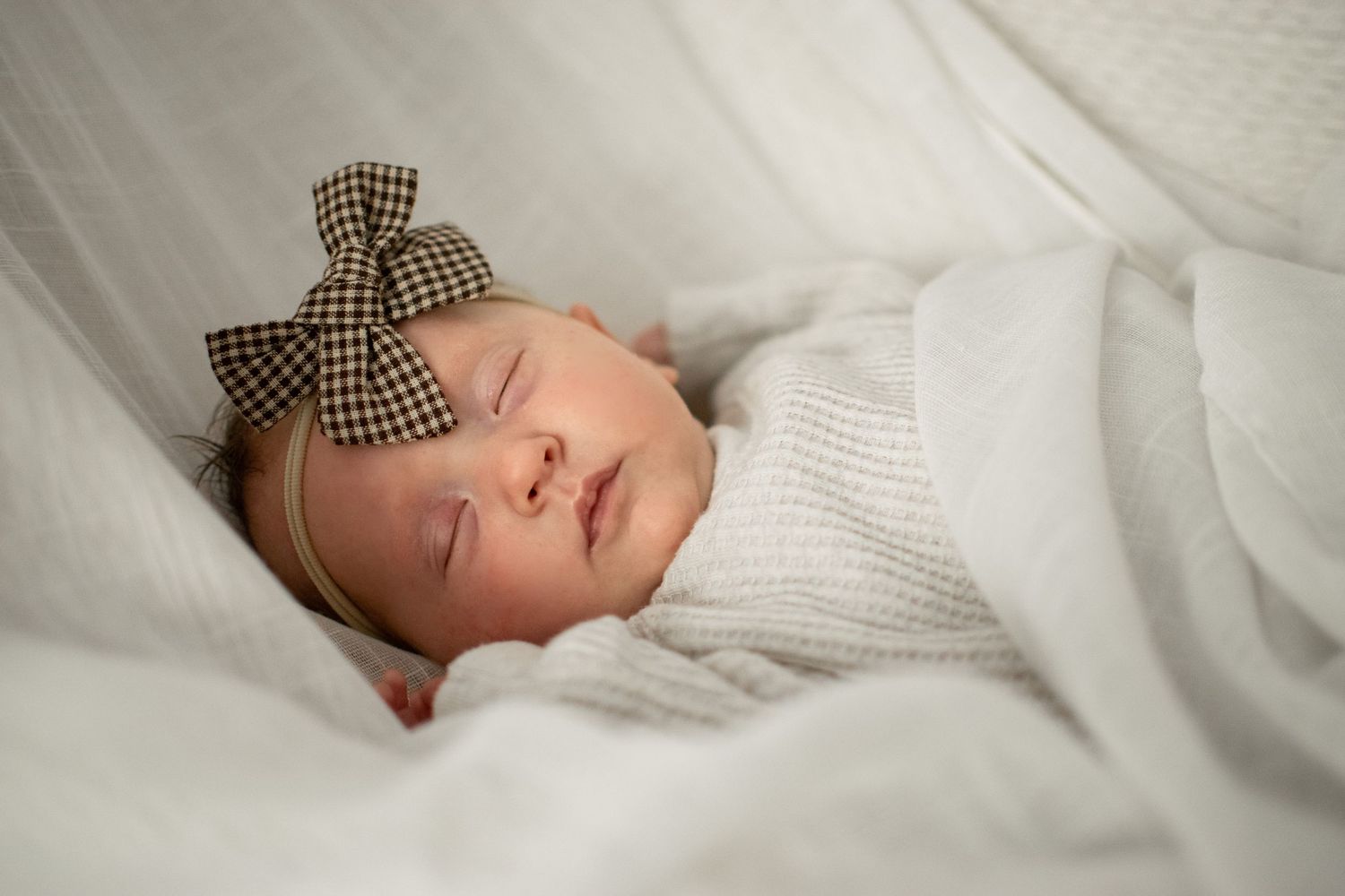 Infant with checkered bow resting on white knit blanket in natural light.