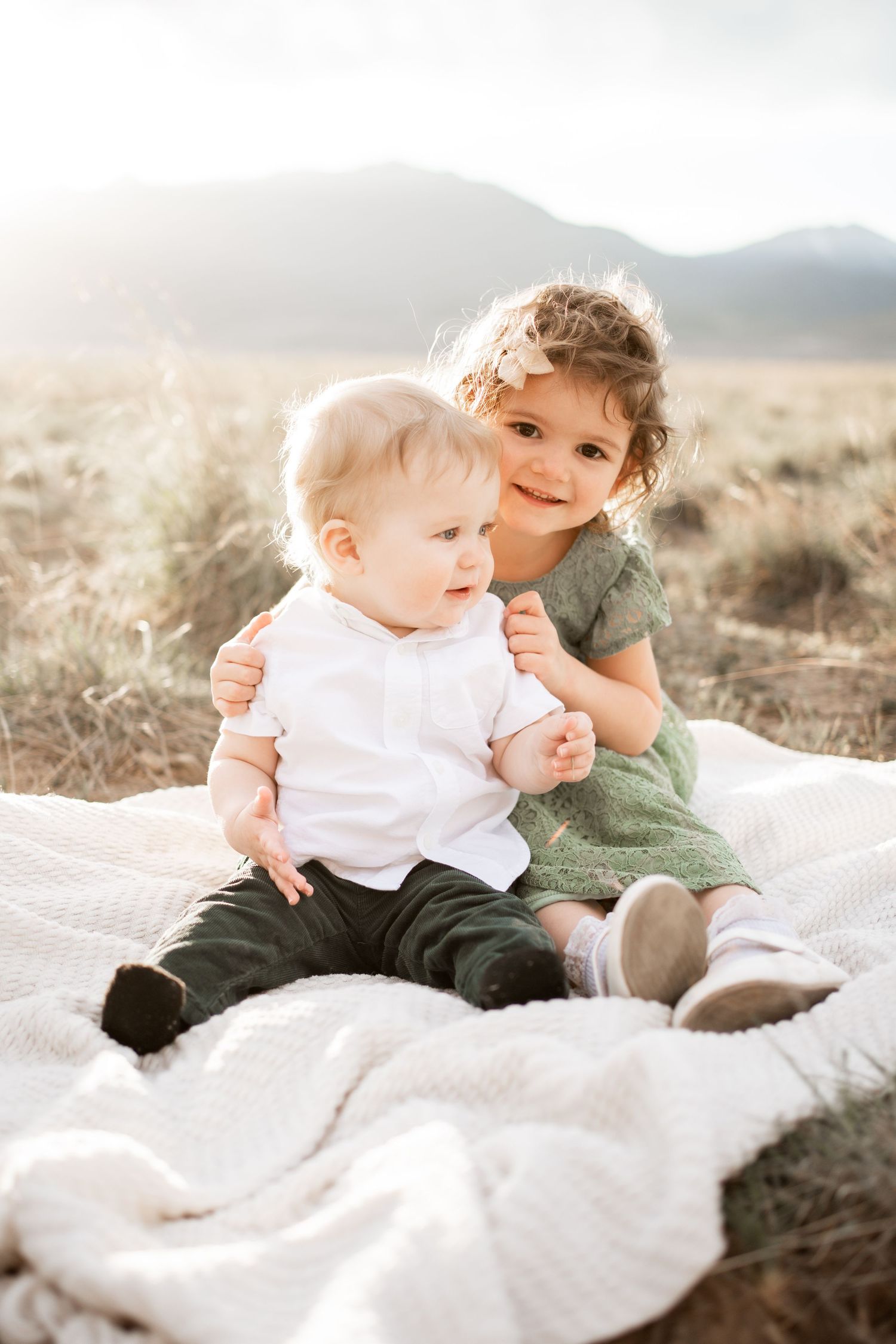 Two young siblings sitting on a blanket outdoors, older girl in green dress hugging baby brother in white shirt.