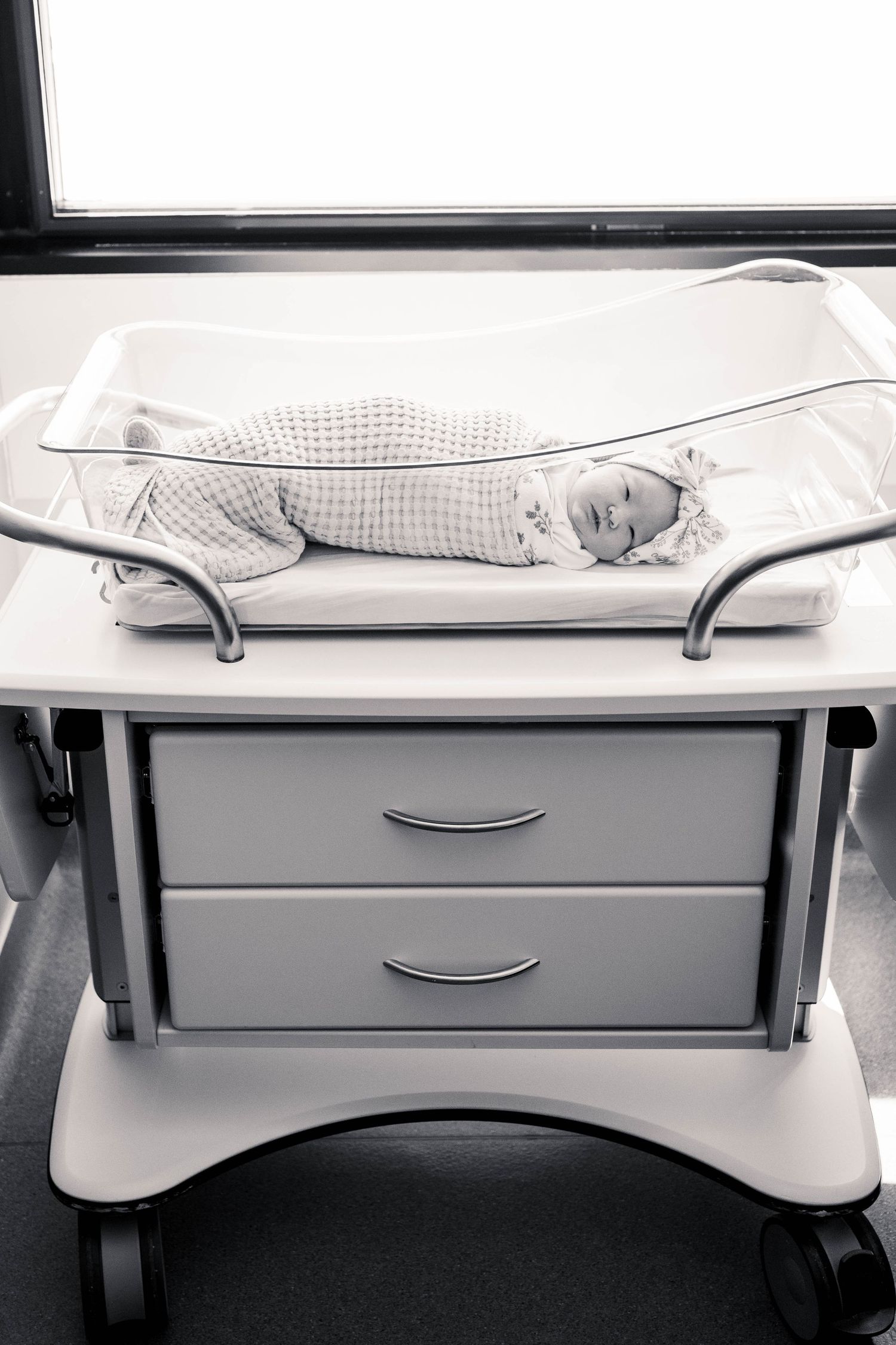Black and white photo of a newborn baby sleeping peacefully in a hospital bassinet on a wheeled cart.