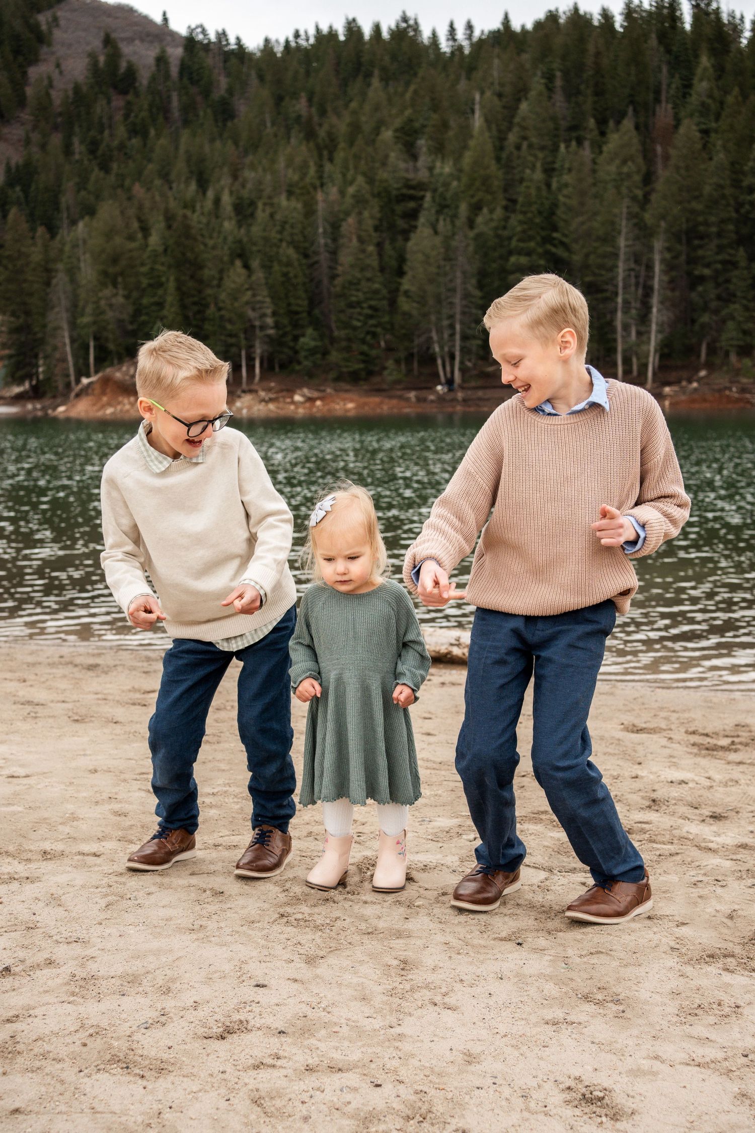Three siblings in cozy sweaters play together on a beach by a mountain lake surrounded by evergreen forest.