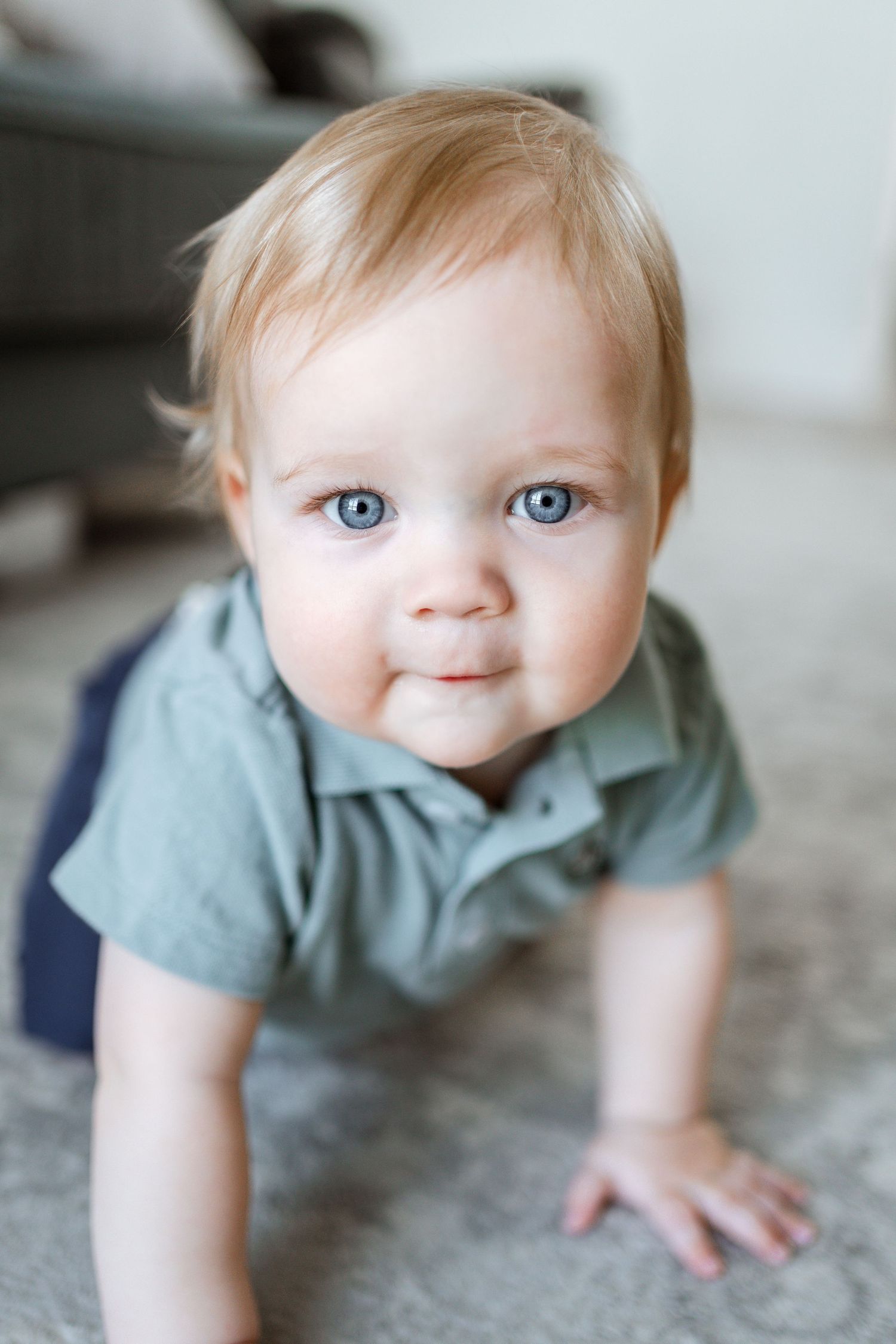 Baby wearing blue shirt crawls on light colored floor with bright eyes and sweet expression.