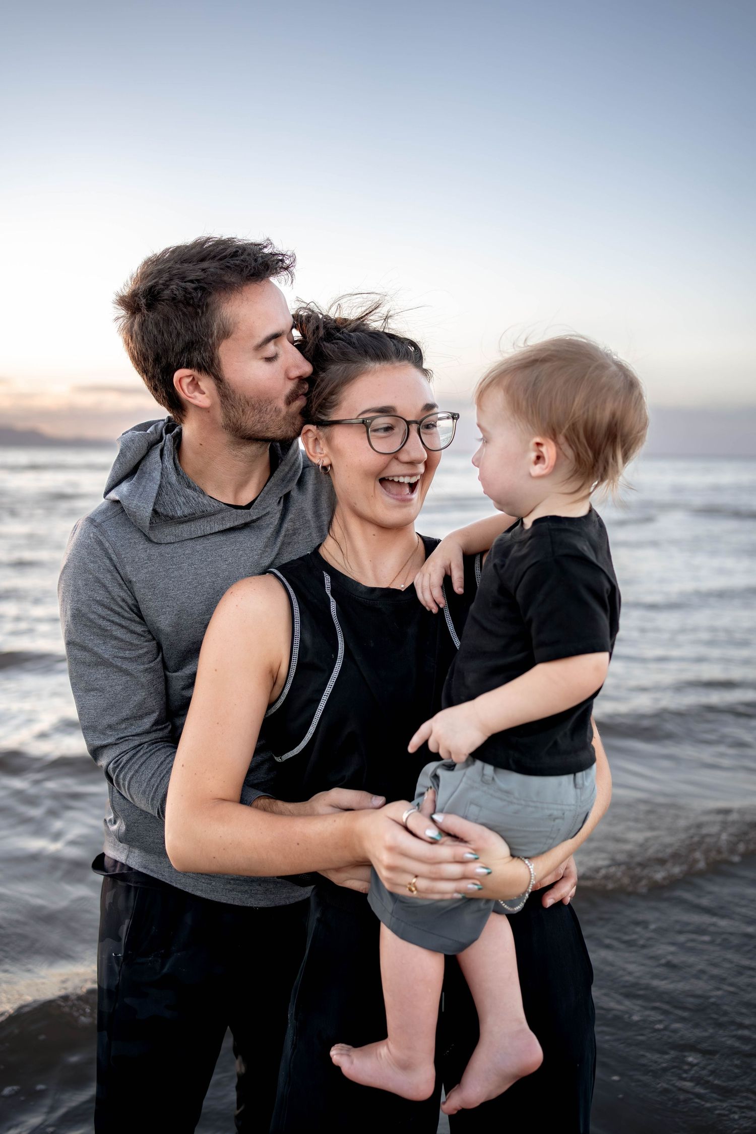 A happy young family with a baby enjoys a sunset beach moment, with dad kissing mom on the cheek.