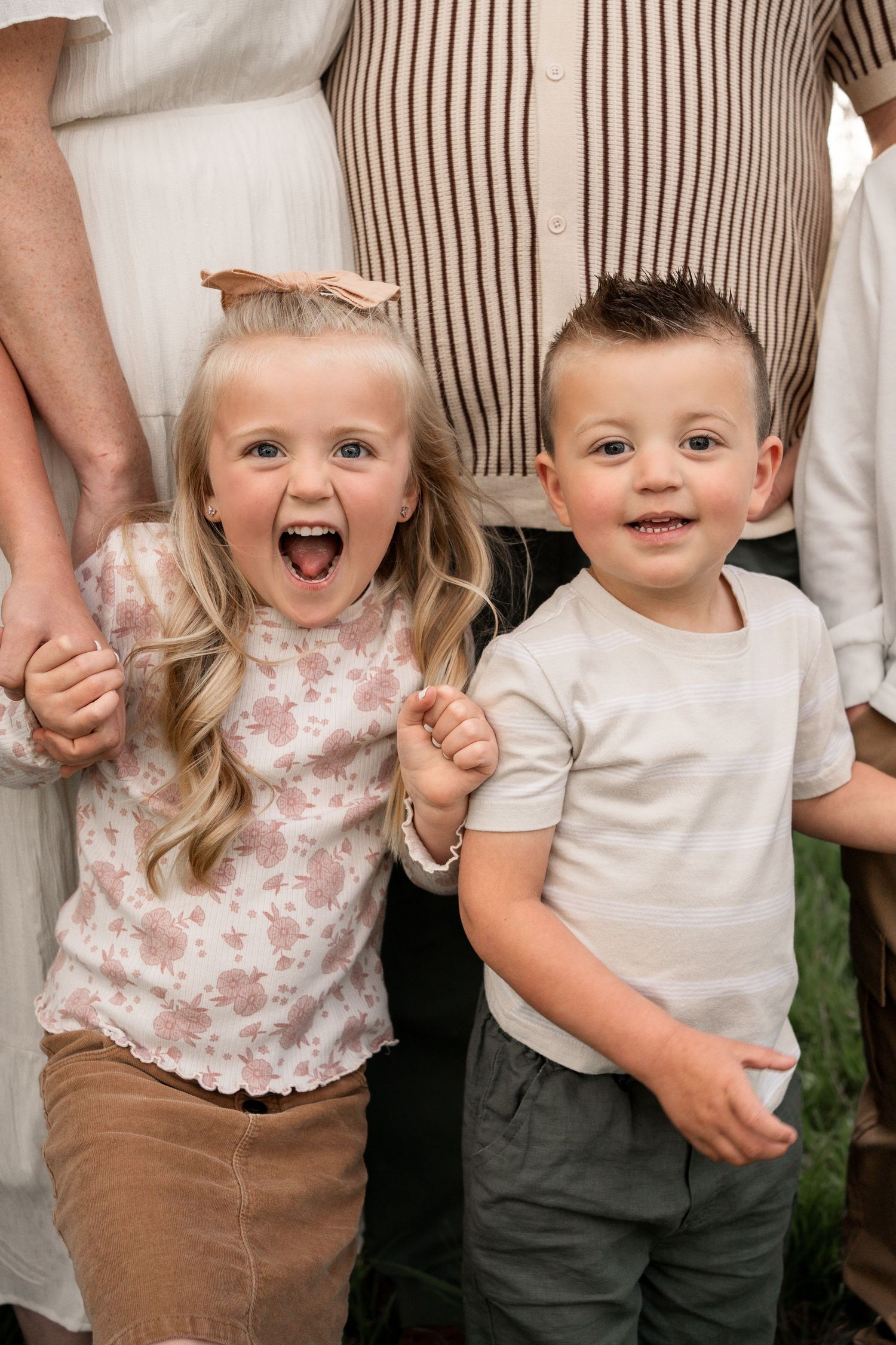Two young siblings smile and laugh together, a blonde girl and dark-haired boy, standing in front of adults outdoors.