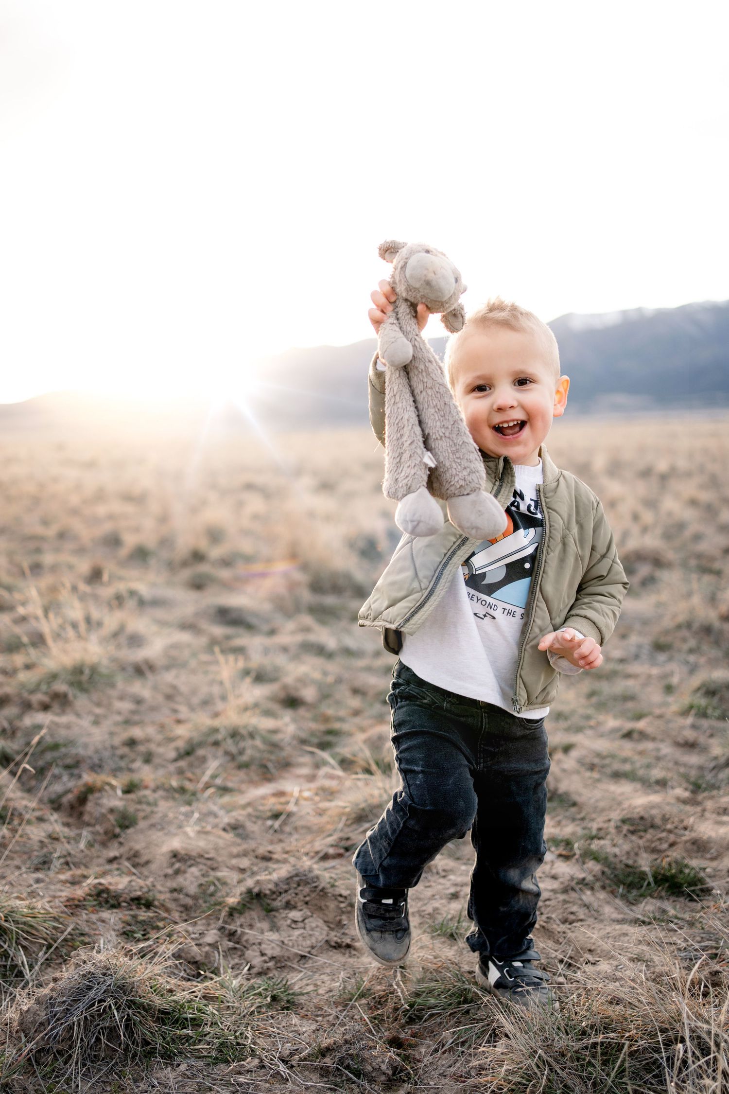 A joyful child runs through a dry field at sunset while holding up a stuffed toy animal.