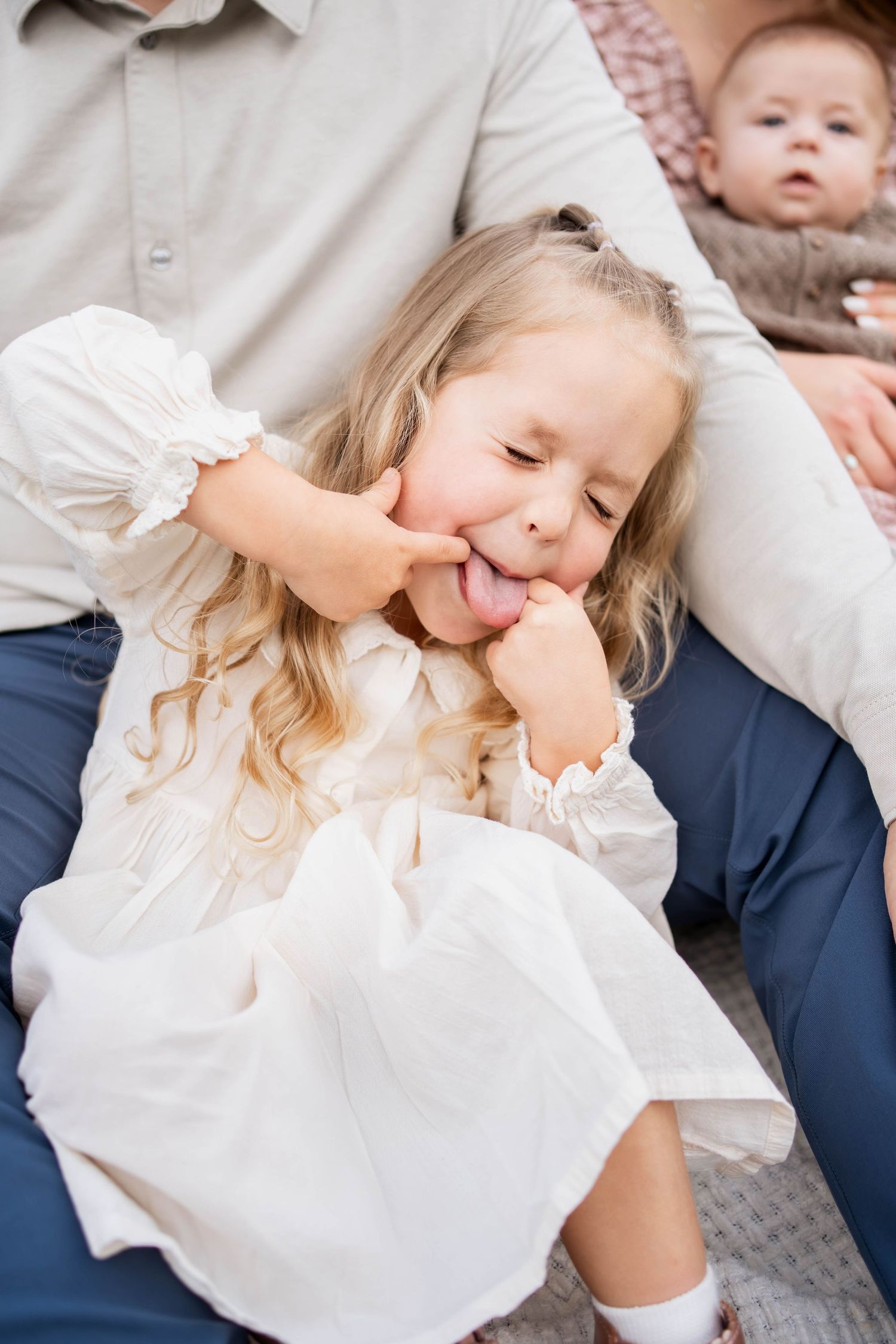 Young girl in a white dress sits on an adult's lap, playfully sticking out her tongue while a baby looks on in the background.