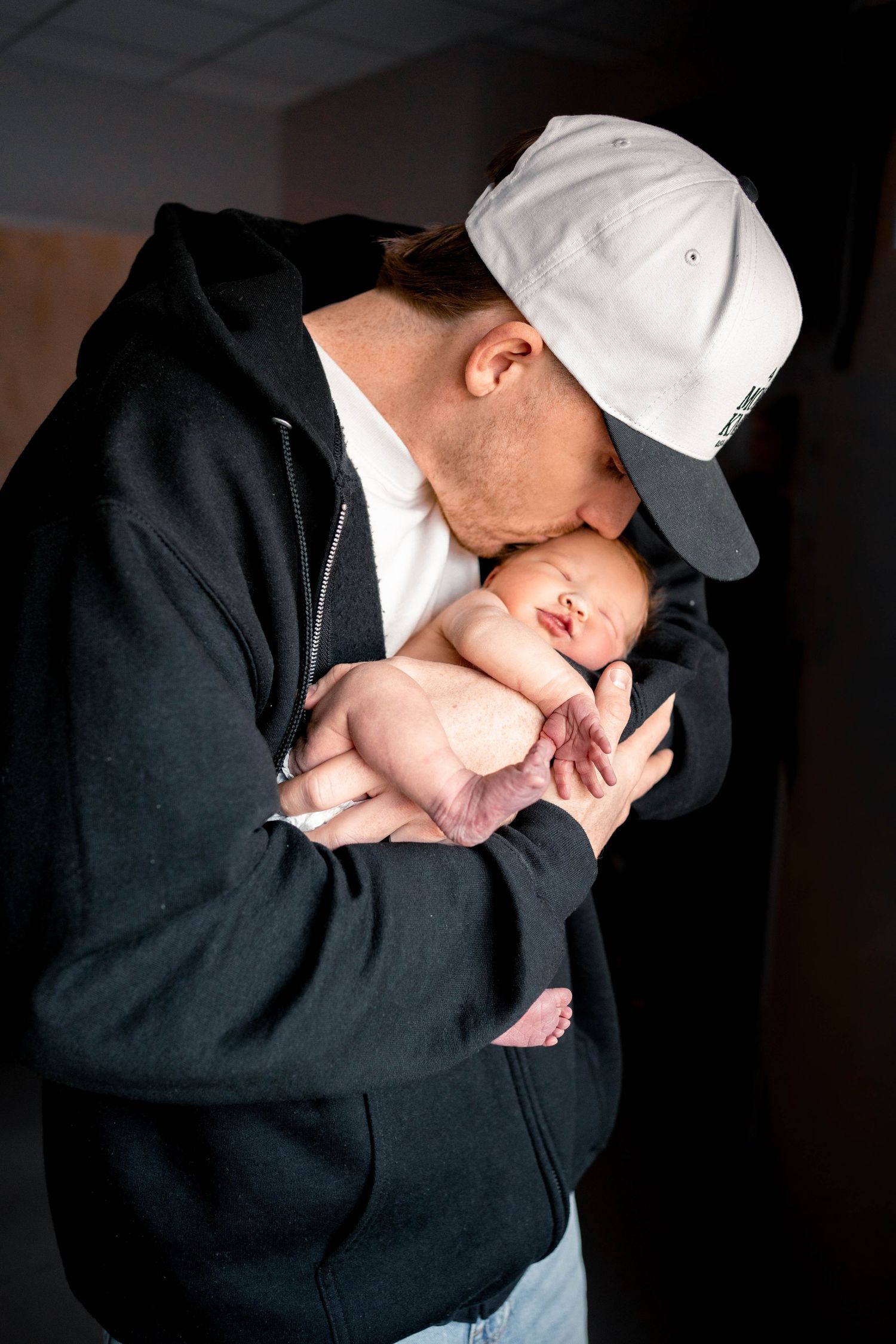 A father in a white cap and black hoodie tenderly kisses his sleeping newborn baby cradled in his hands.