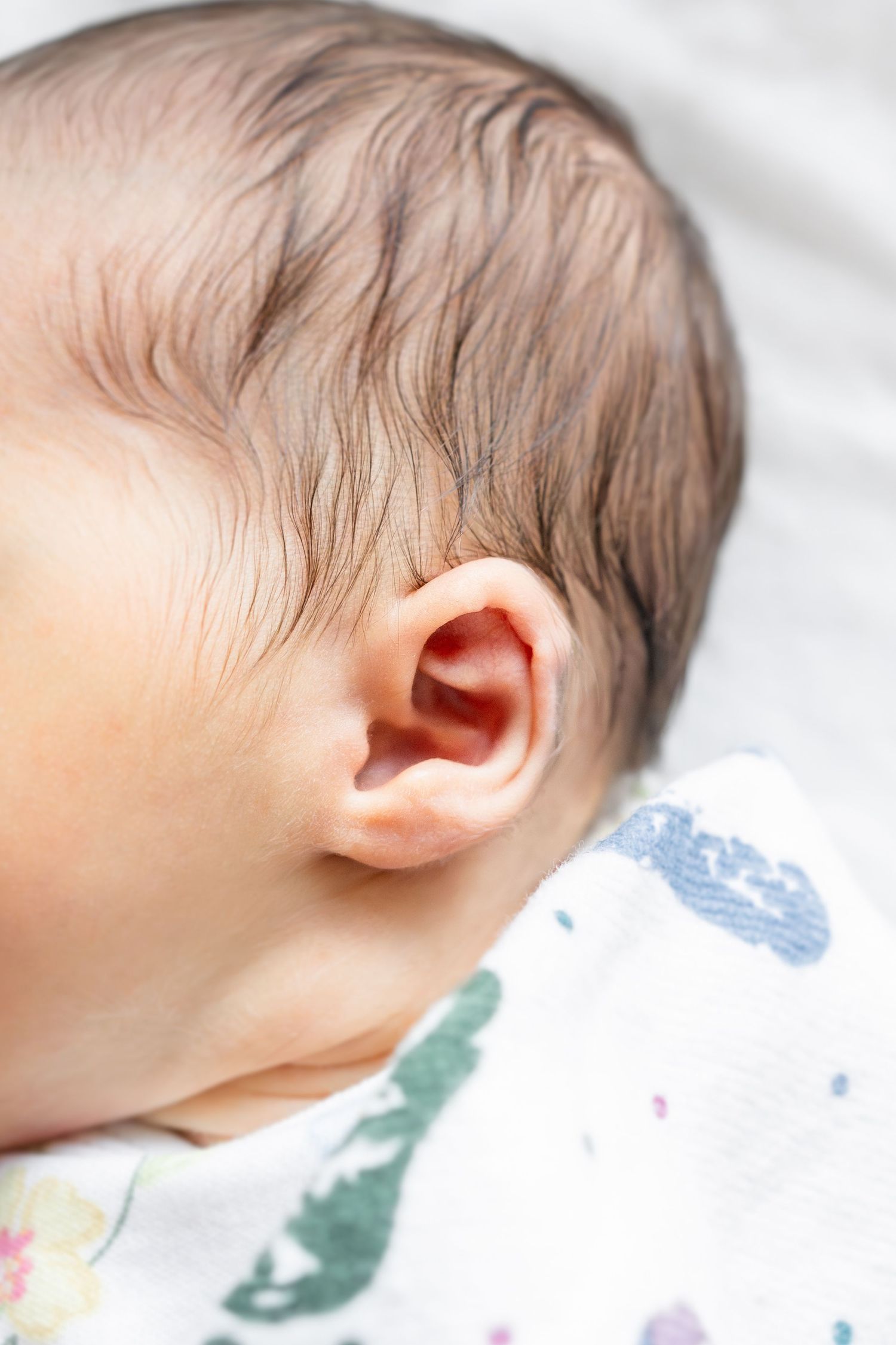 Close-up of a newborn baby's ear and dark hair, lying on a white patterned blanket.