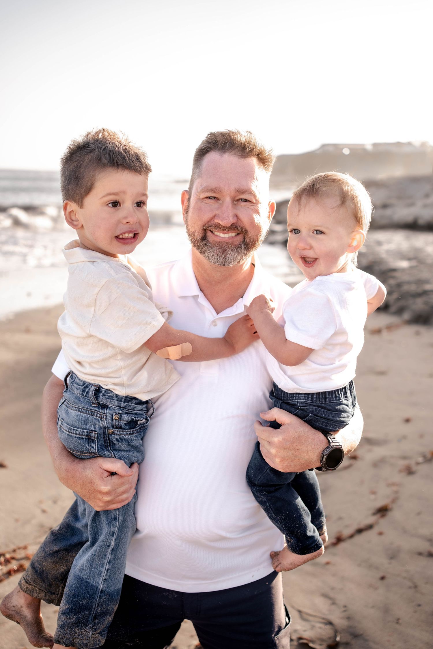 A smiling father holds two young boys on a sunny beach, all wearing white shirts and jeans.