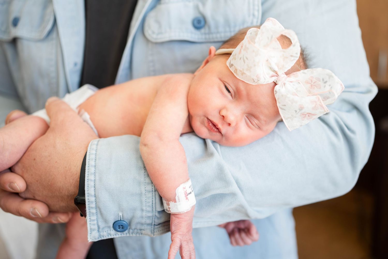 Newborn baby girl sleeping in parents arms wearing a white floral bow headband.