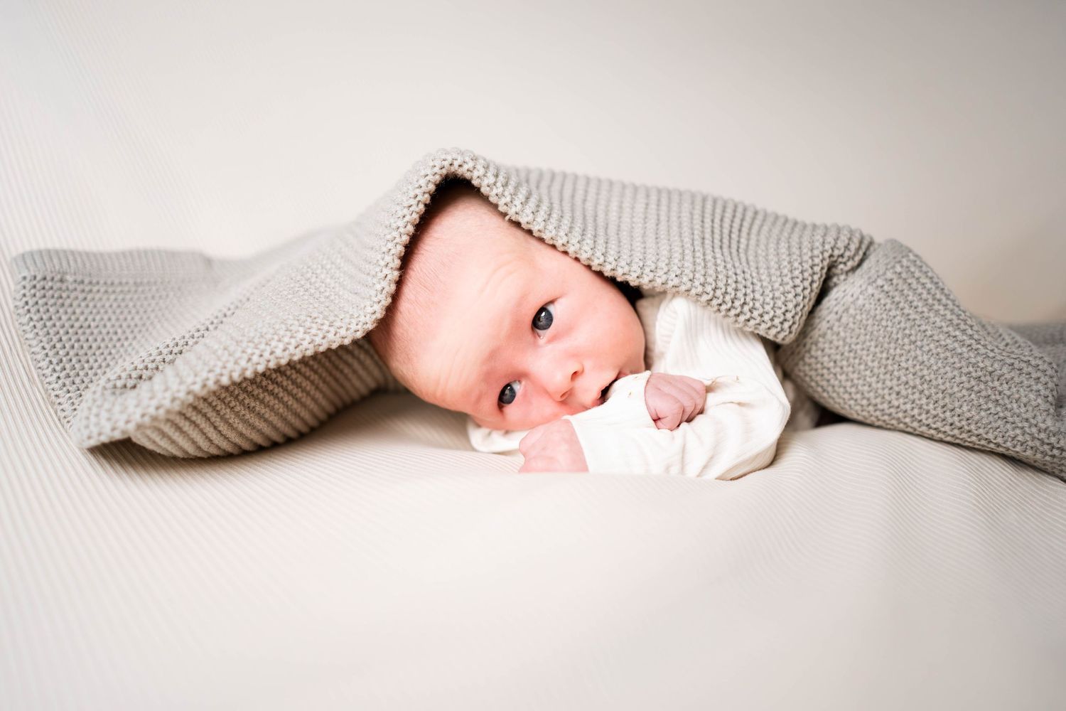 Newborn baby peeking out from under a soft gray knit blanket, lying on a white surface.