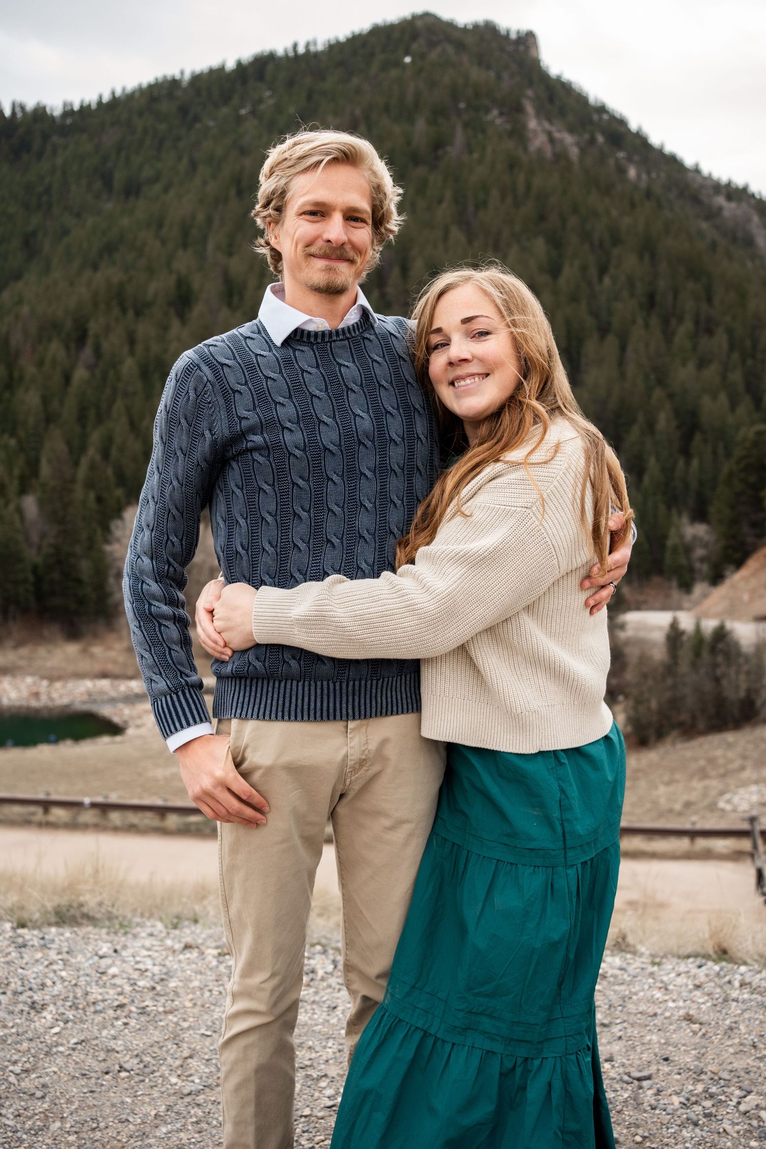 A couple embraces while standing on a rocky beach with evergreen-covered mountains and cloudy skies behind them.