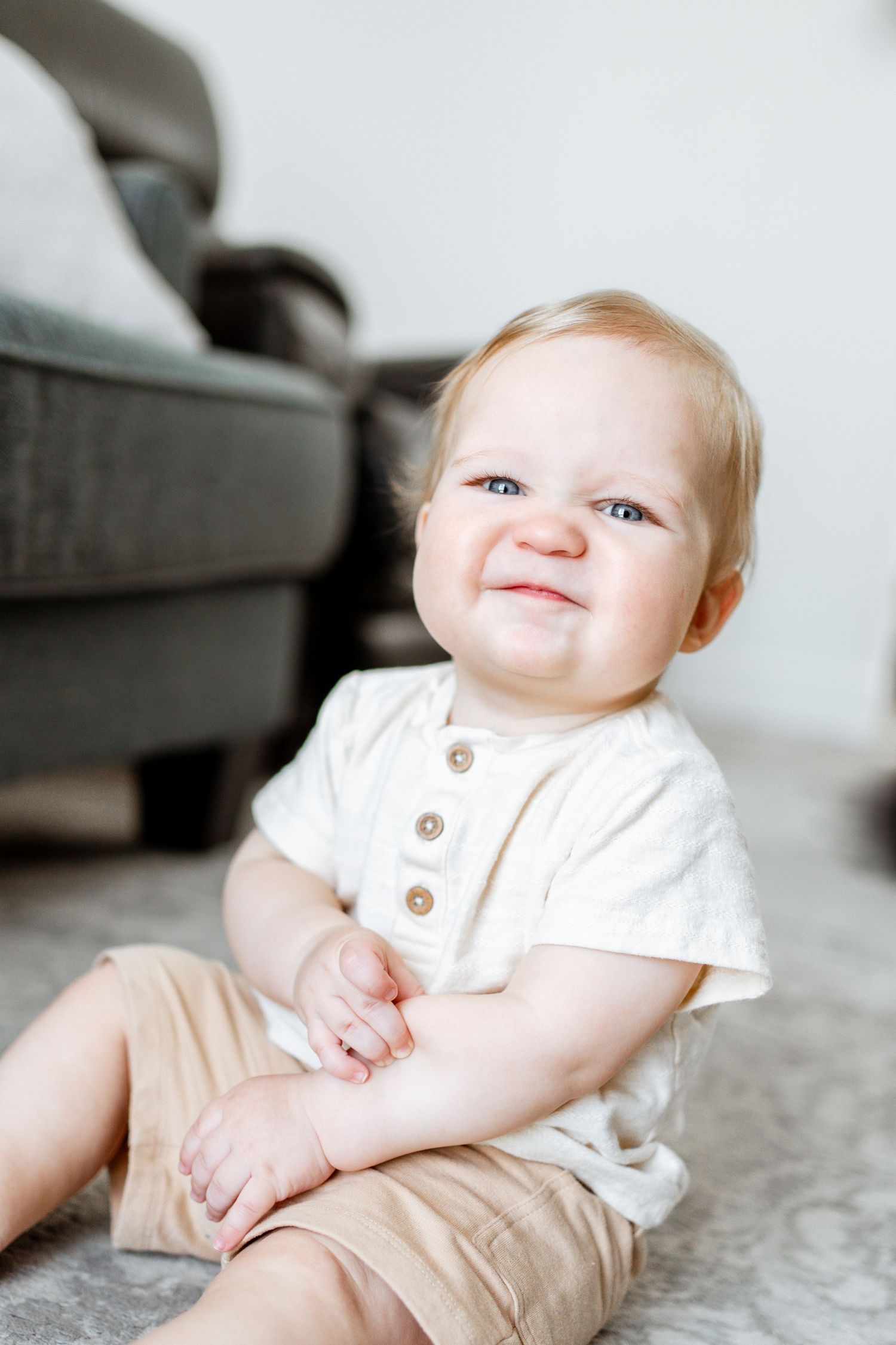 A baby in a white shirt and khaki pants sits on a carpet while smiling at the camera.
