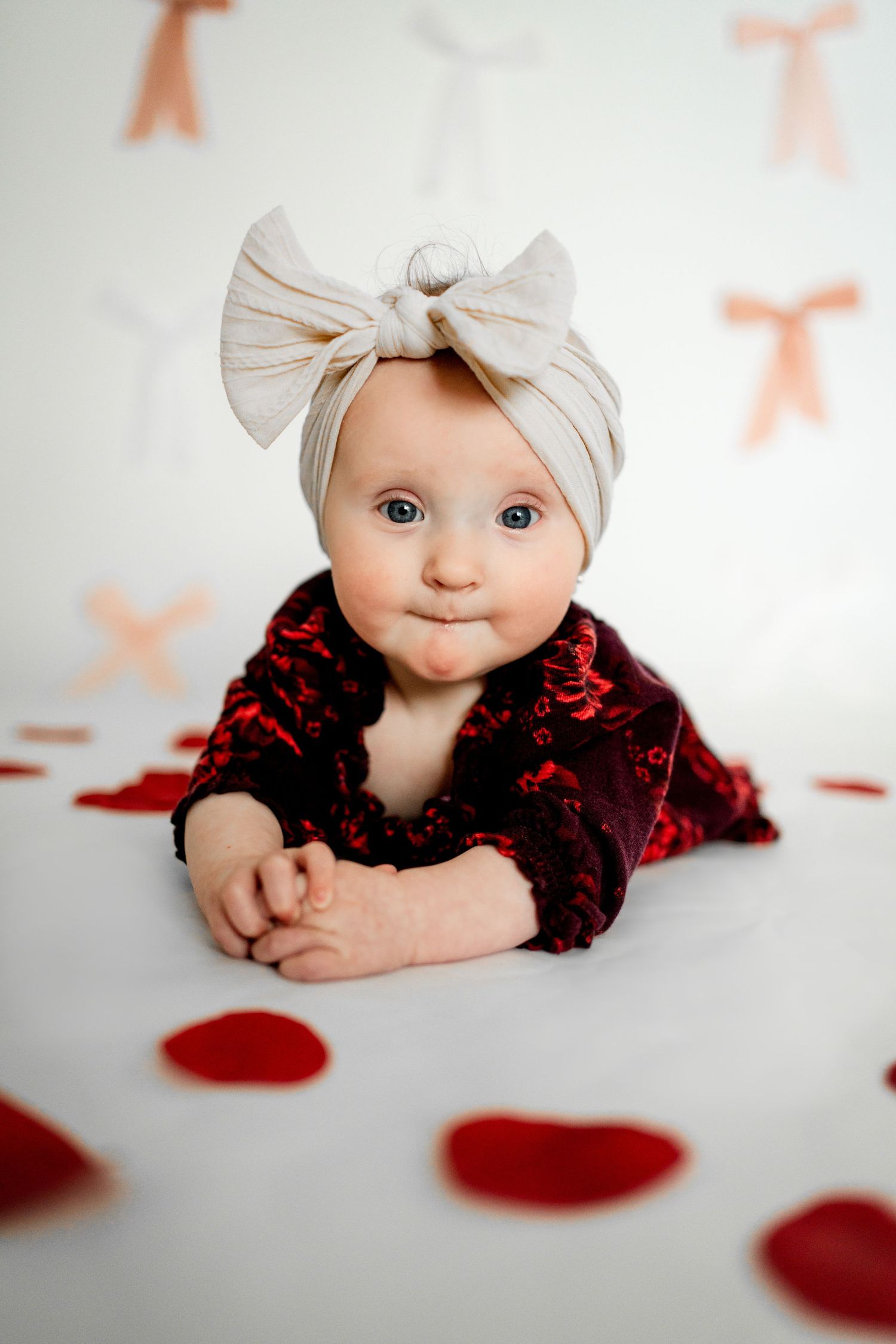 Baby wearing white bow headband and burgundy lace outfit lies on white surface with red heart decorations.